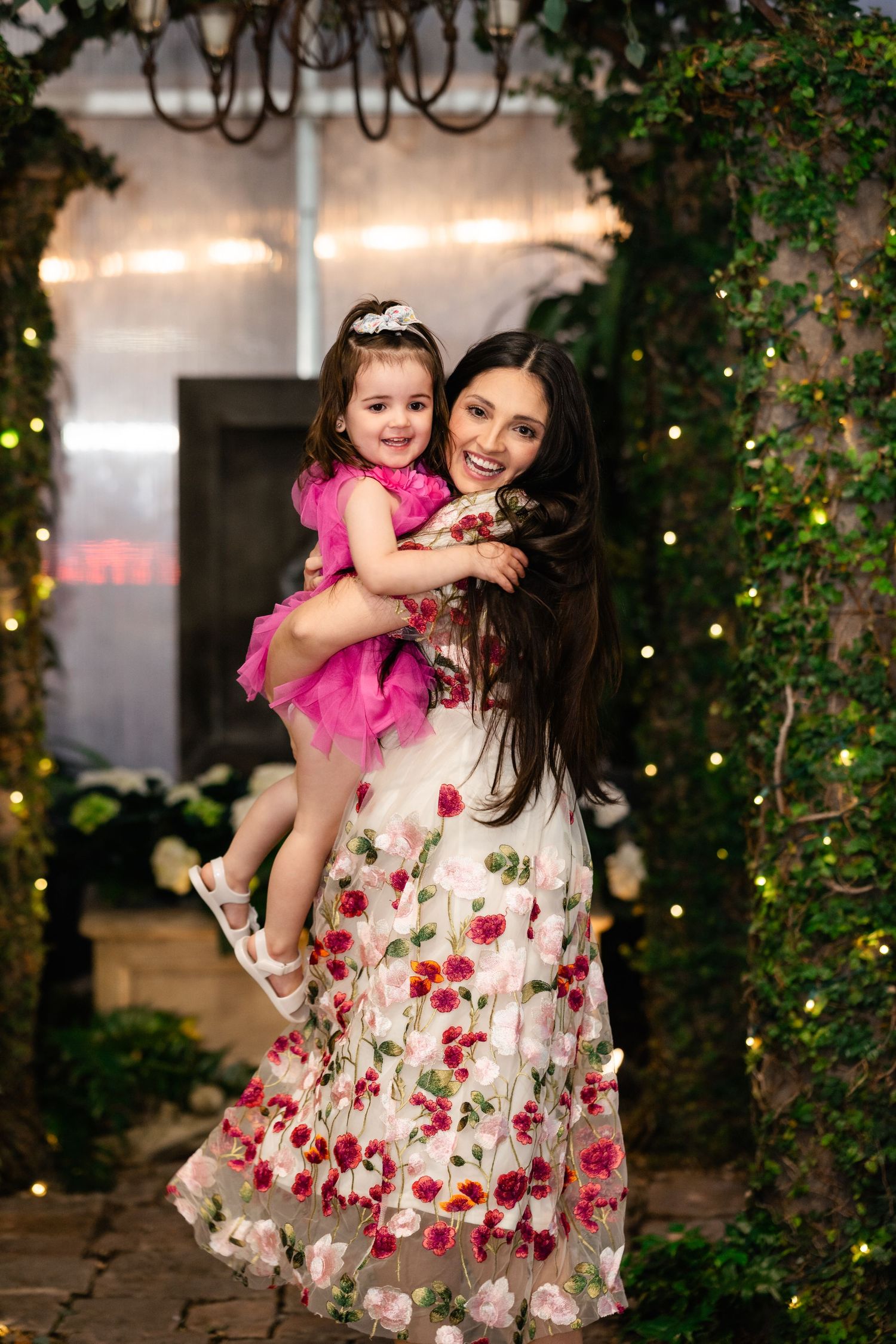 A mother and daughter in coordinating pink and floral dresses pose together in a garden setting with twinkle lights.