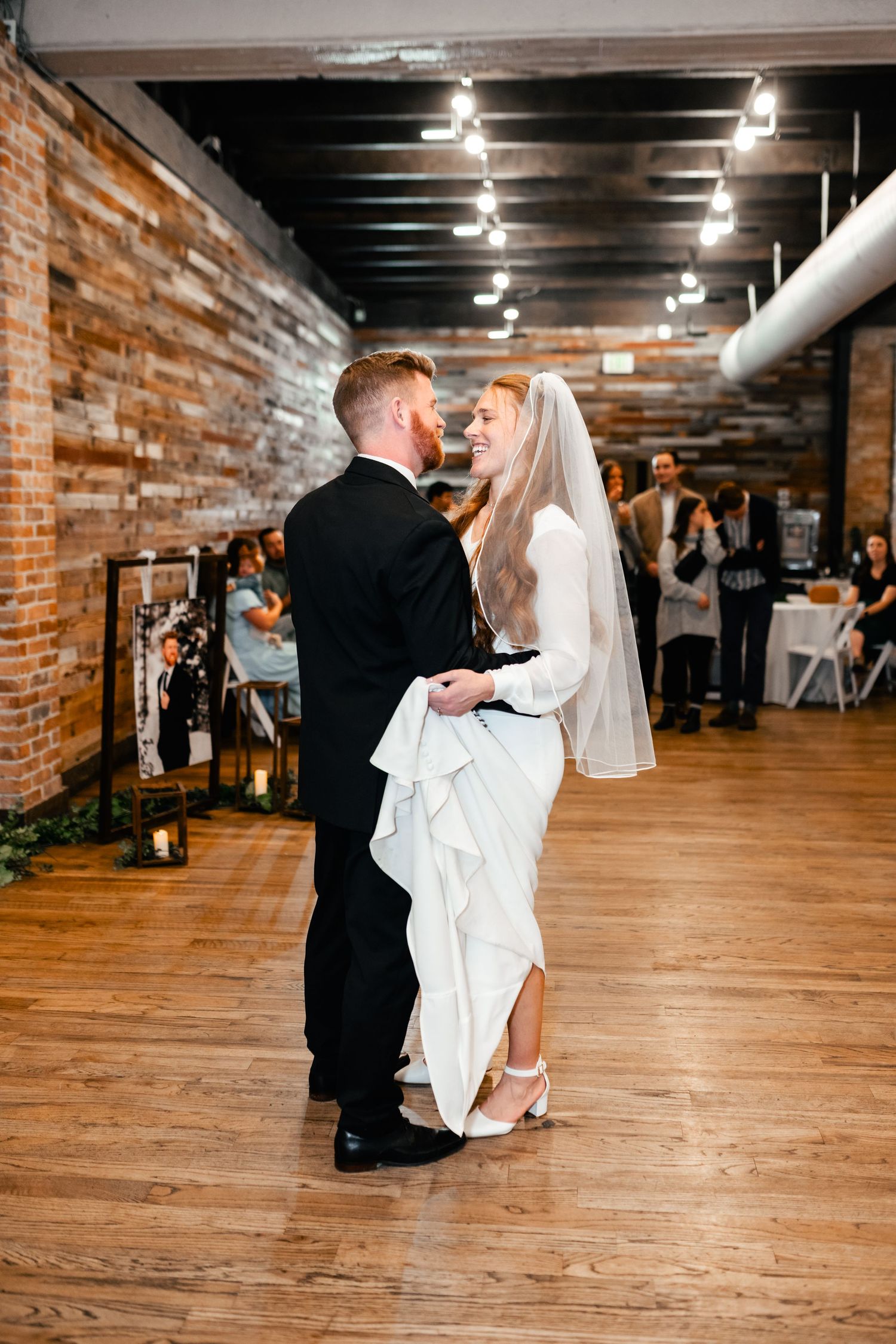 A couple shares their first dance at a rustic wedding reception with exposed brick walls and hardwood floors.