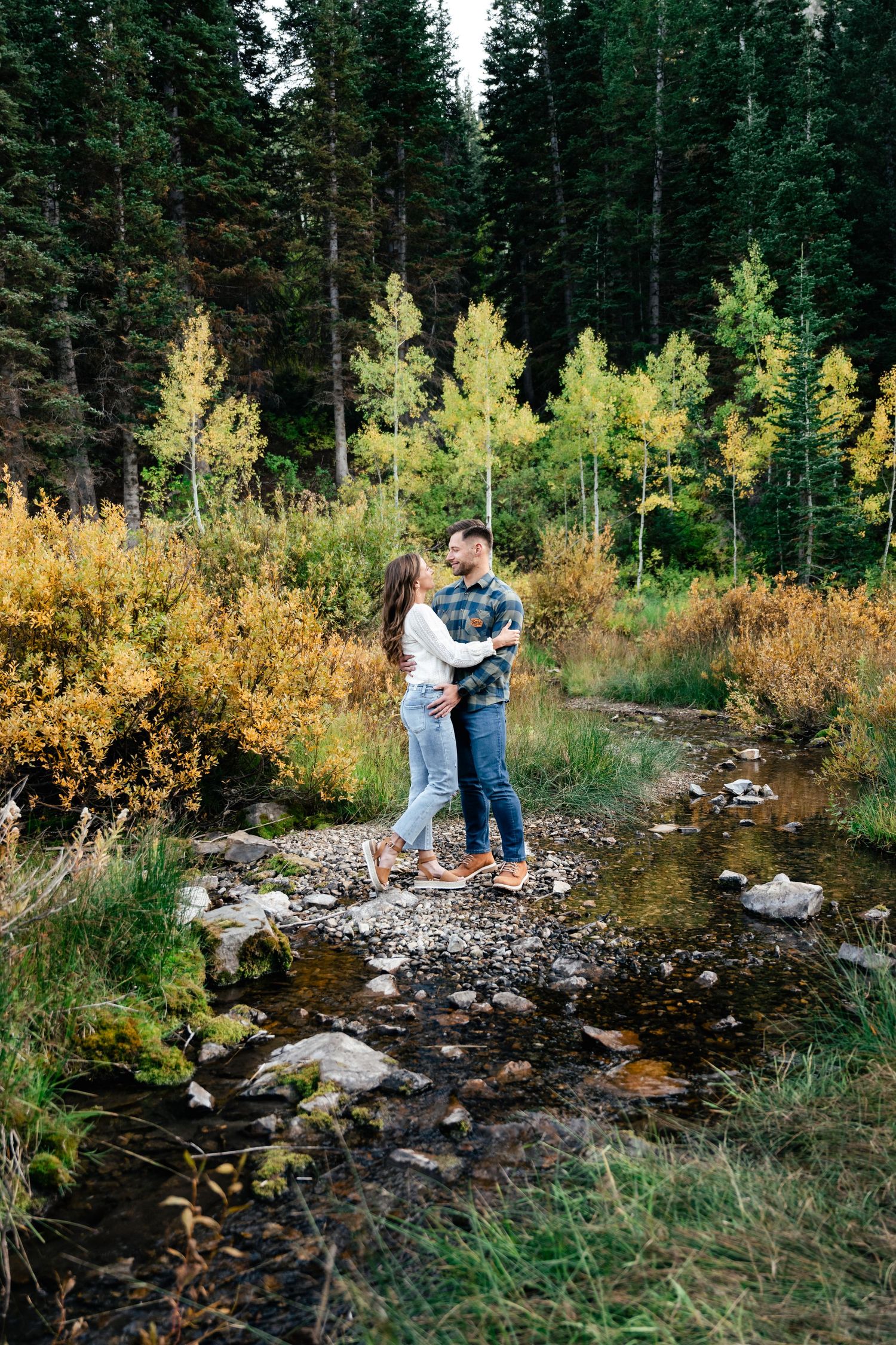 A couple embraces on rocky creek bed surrounded by autumn foliage and evergreen forest.