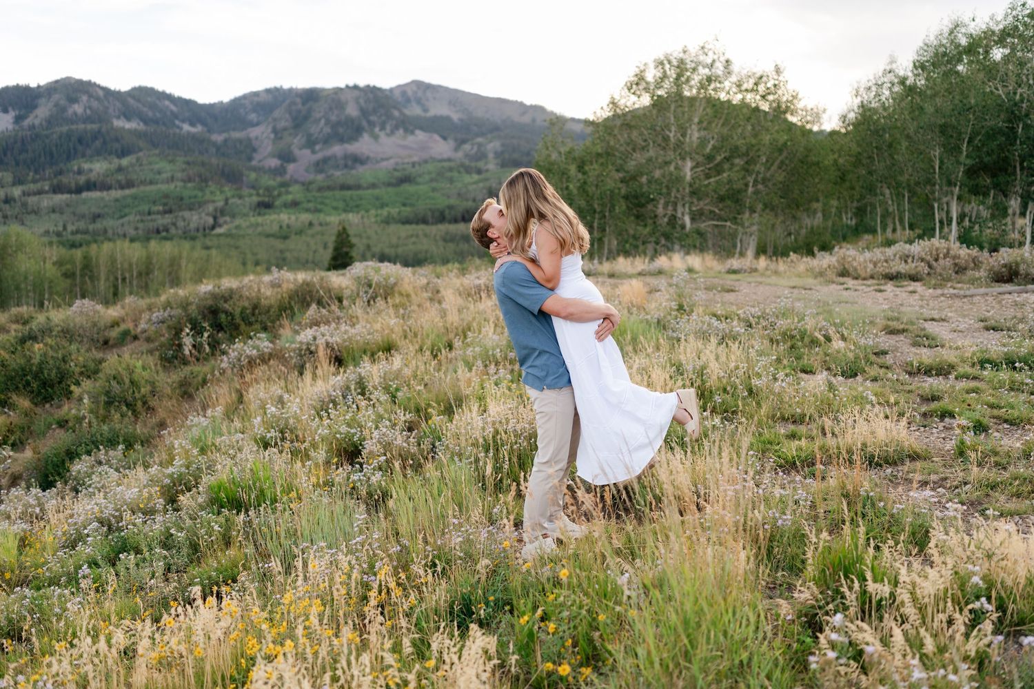Mountain engagement photos near Park City. Couple embracing in a mountain meadow, man lifting woman with scenic hills backdrop.