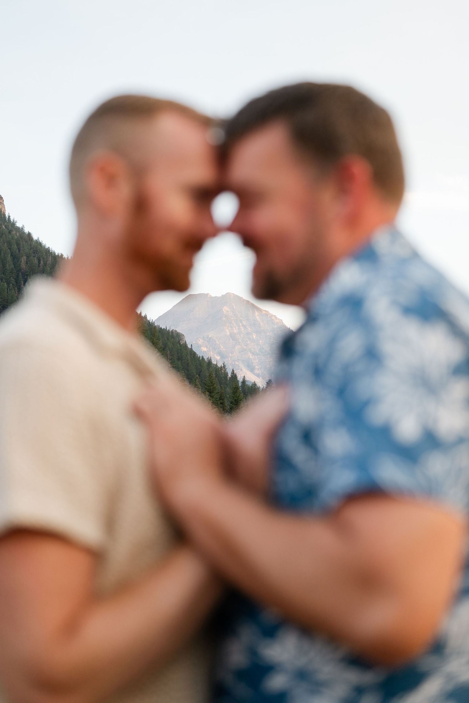 Two men touch foreheads intimately with a scenic mountain and pine forest backdrop in soft focus.