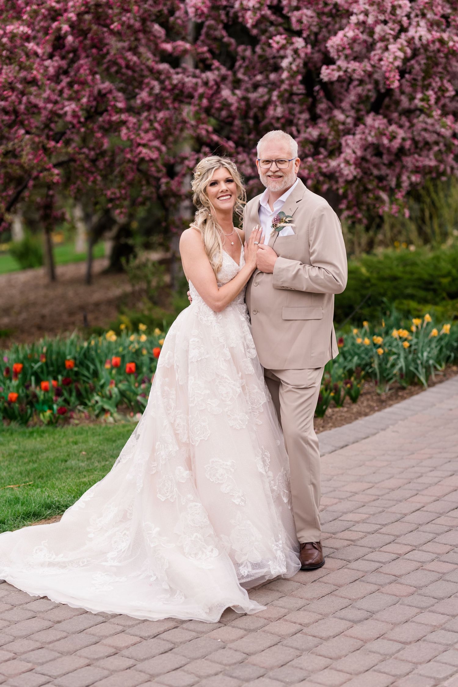 A couple poses together on a brick pathway with blooming pink cherry blossoms and tulips in the garden background.