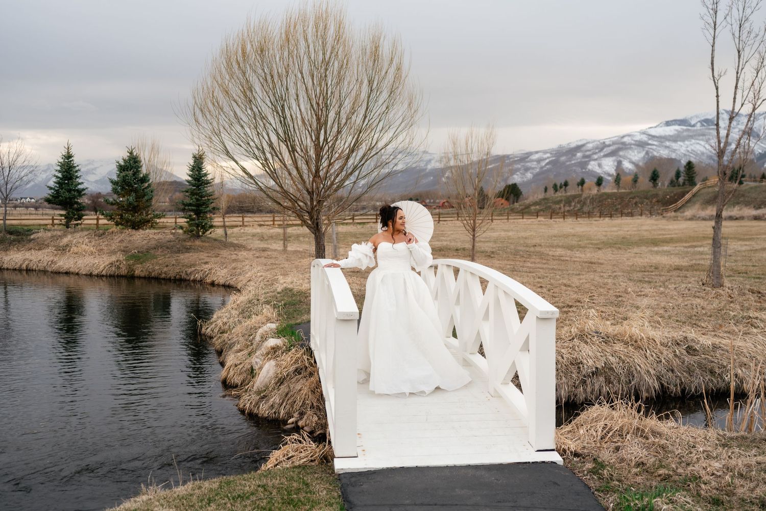 Bride with parisol on a bridge at winter lake scene with bare trees and snowy mountains in the background. River Bottoms Ranch Photographer.