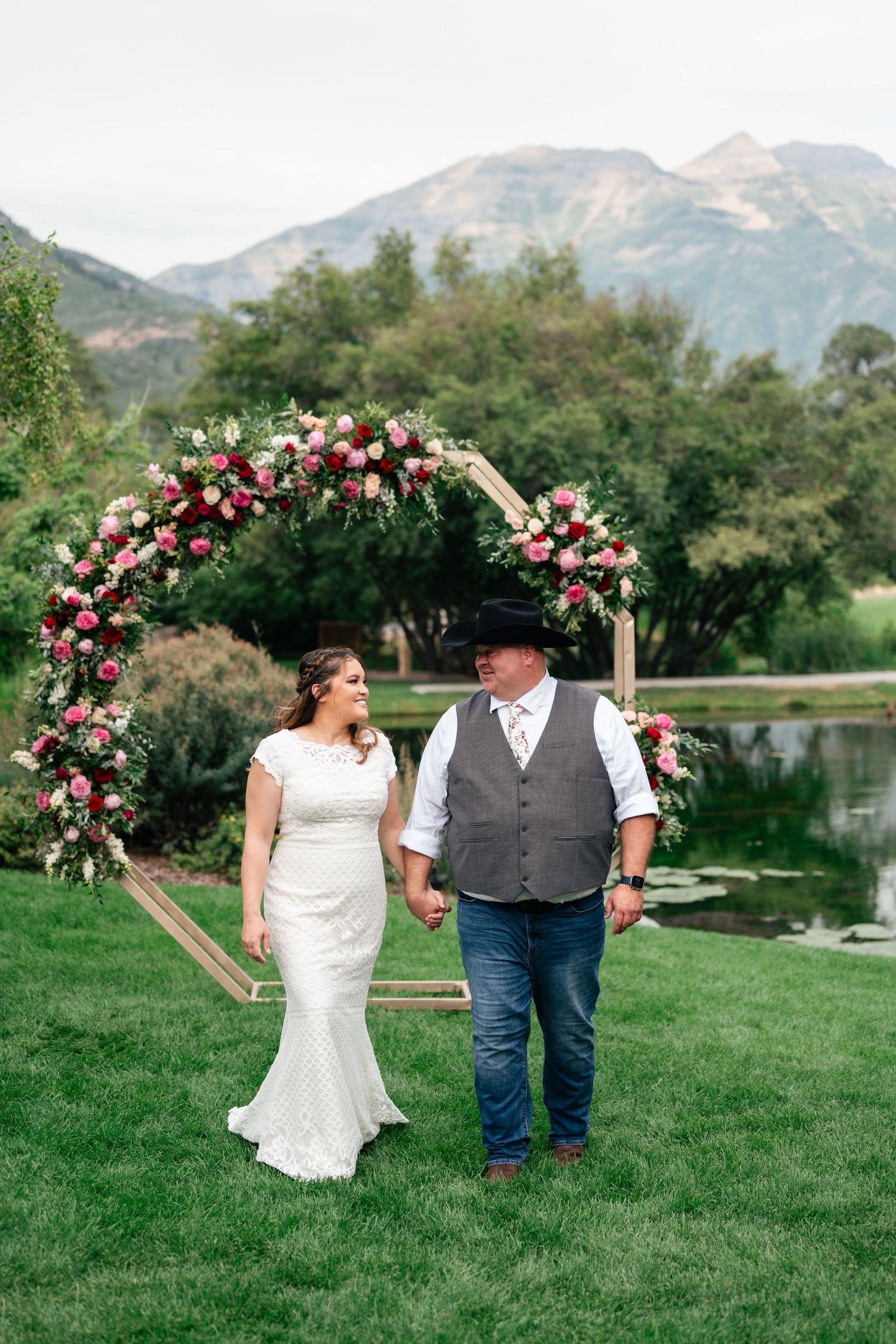 A couple walks together on green grass with mountains and a lake in the background during their rustic outdoor wedding. Conrad Ranch.