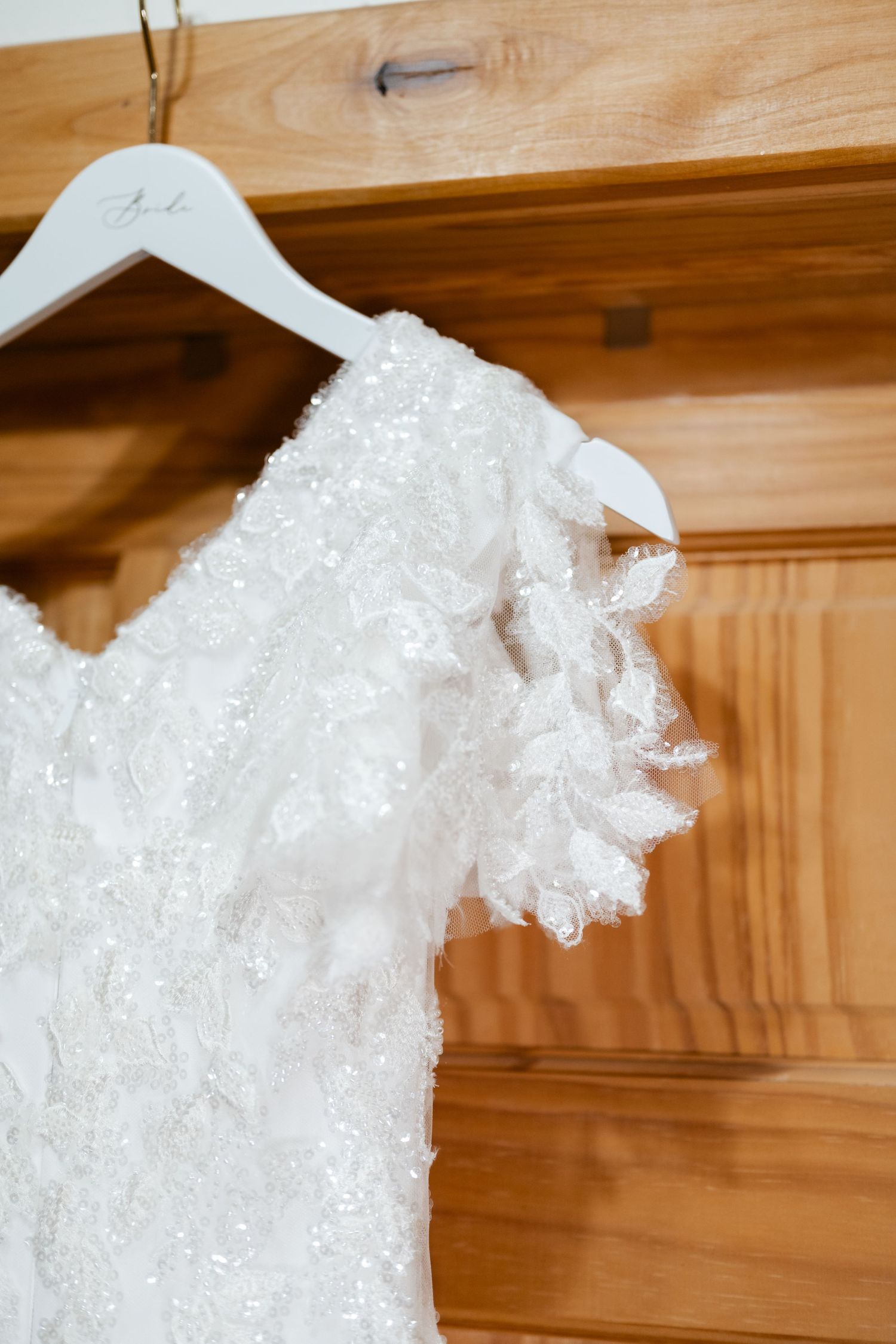 Delicate white lace wedding dress detail hanging on a wooden hanger against a warm wood background.