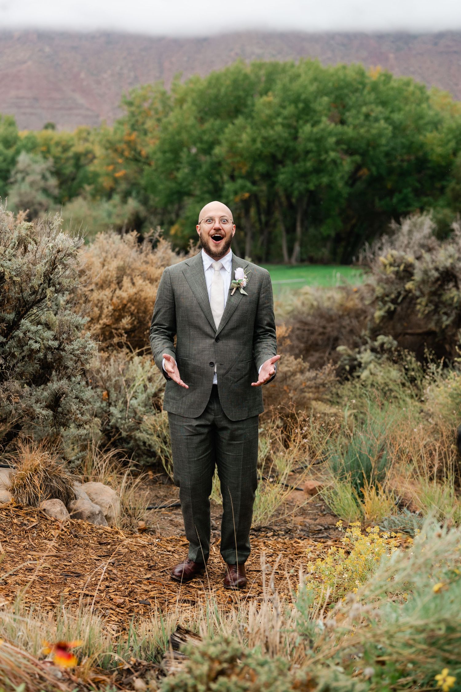 A groom poses cheerfully in a charcoal suit among desert landscaping and fall foliage.