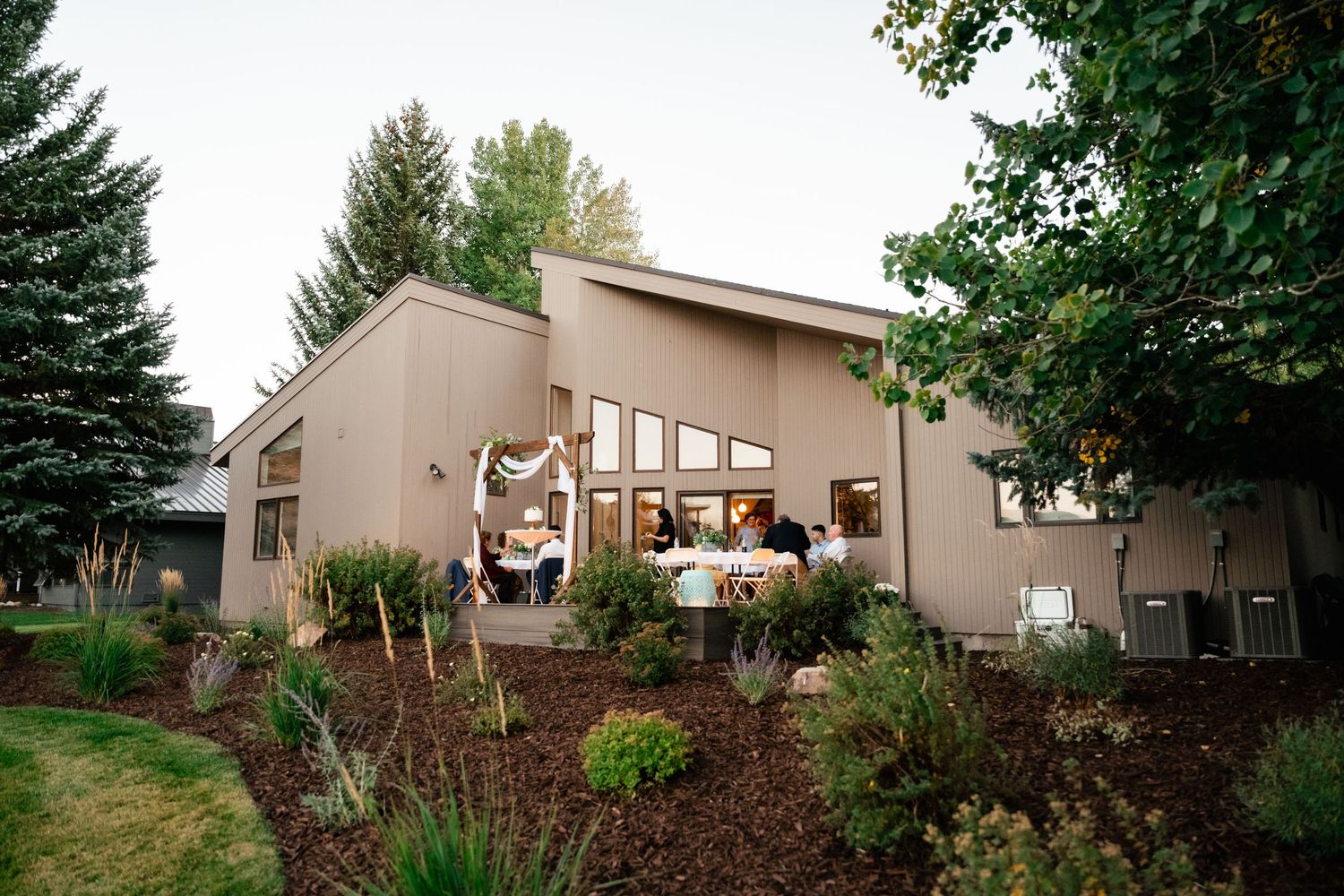 Modern beige house with angled roof and large windows surrounded by evergreen trees.