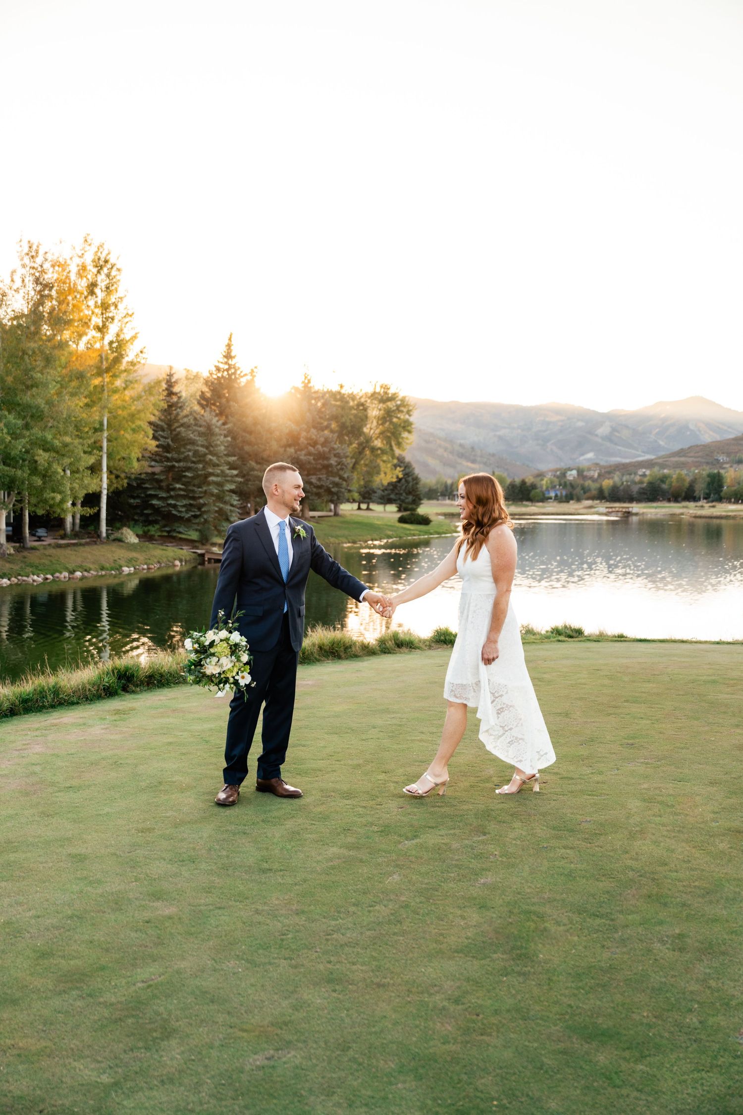 A playful couple dances on grass with a lake and mountains in the background.