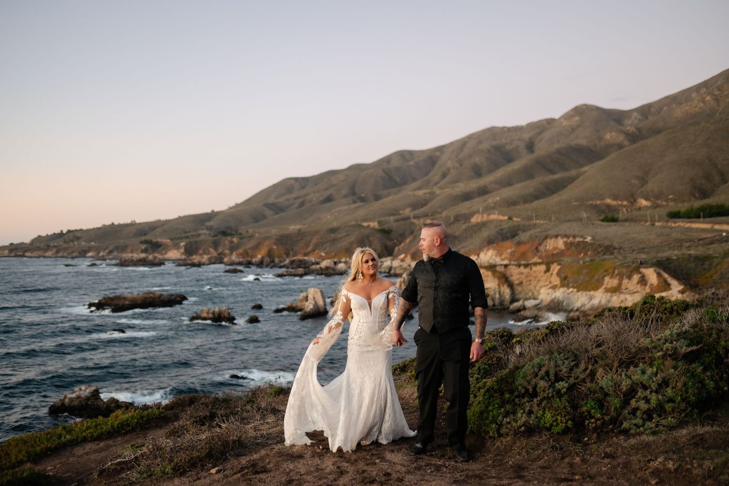 Wedding couple walking along Big Sur coastline with mountains and ocean in background.
