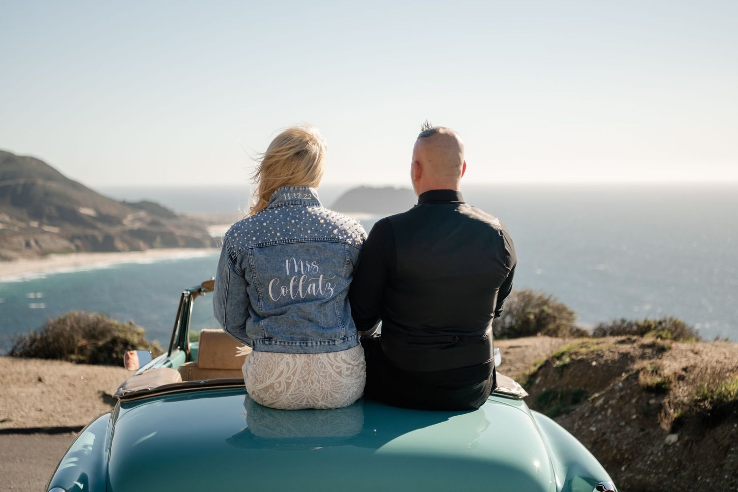 Couple in custom wedding jackets sitting on vintage car overlooking ocean vista.