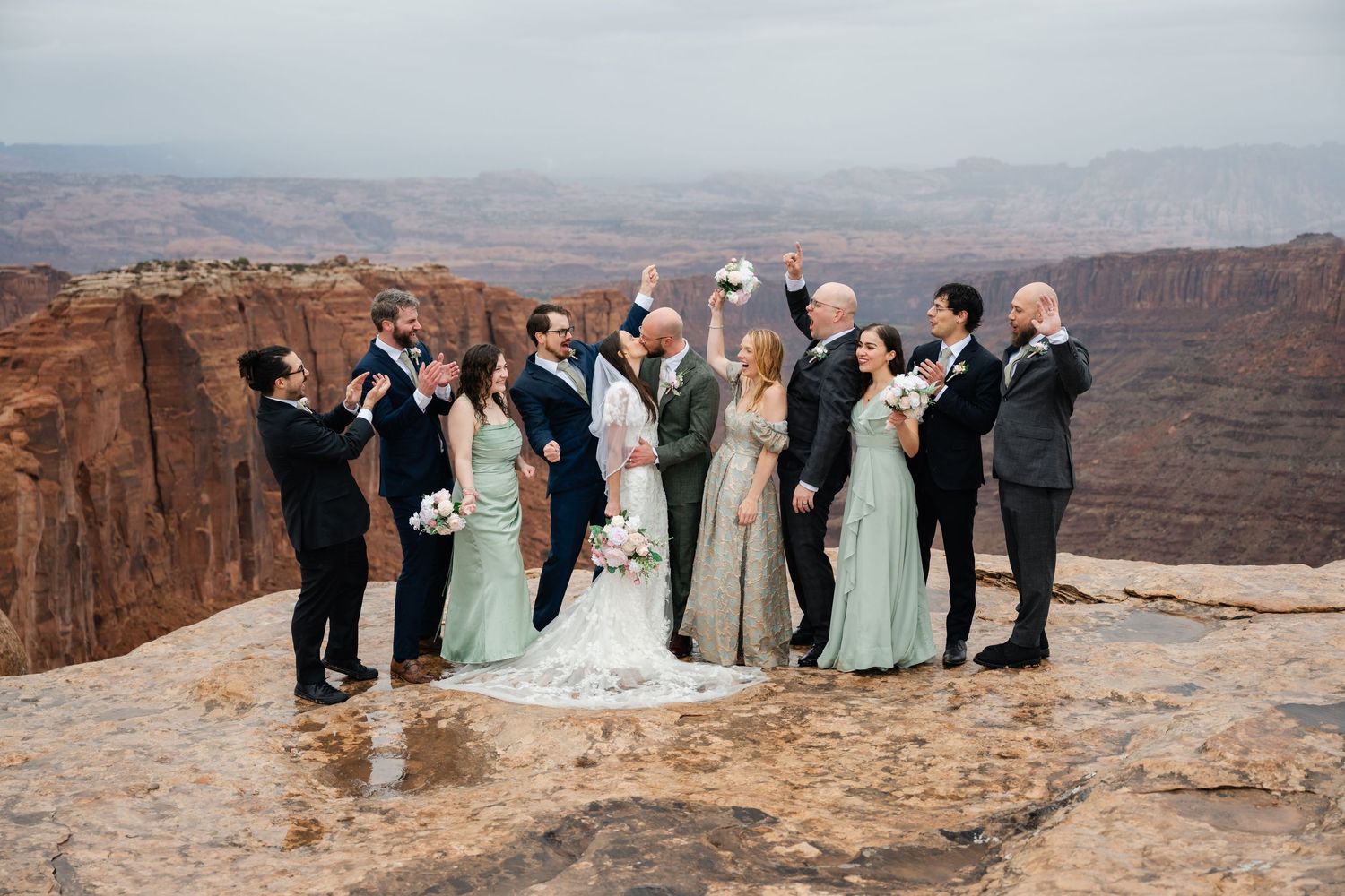 Wedding group poses along scenic canyon overlook with vast desert landscape in background.