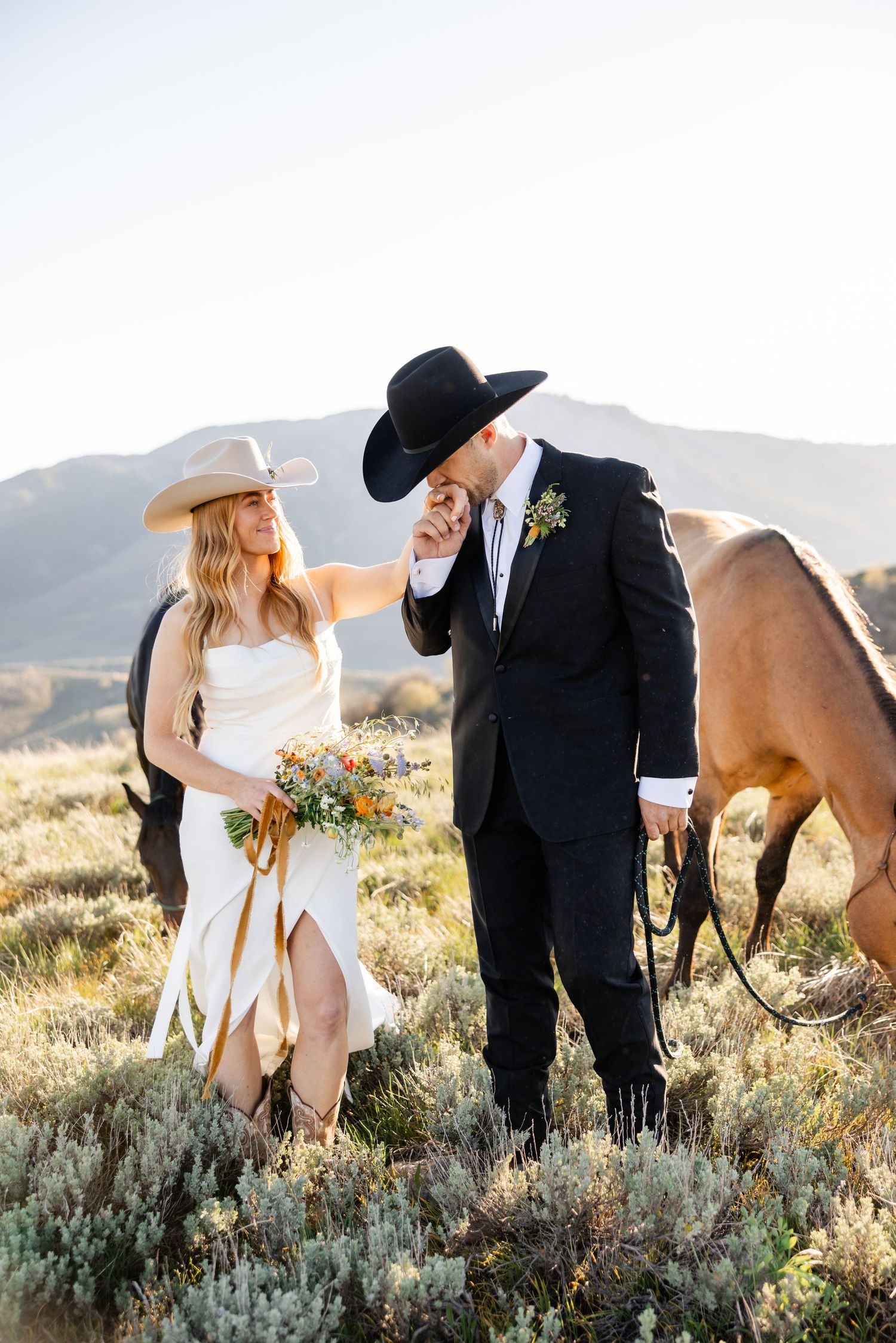 A couple in wedding attire poses together in a desert landscape with wild horses and sagebrush at sunset. Utah Elopement Photographer.