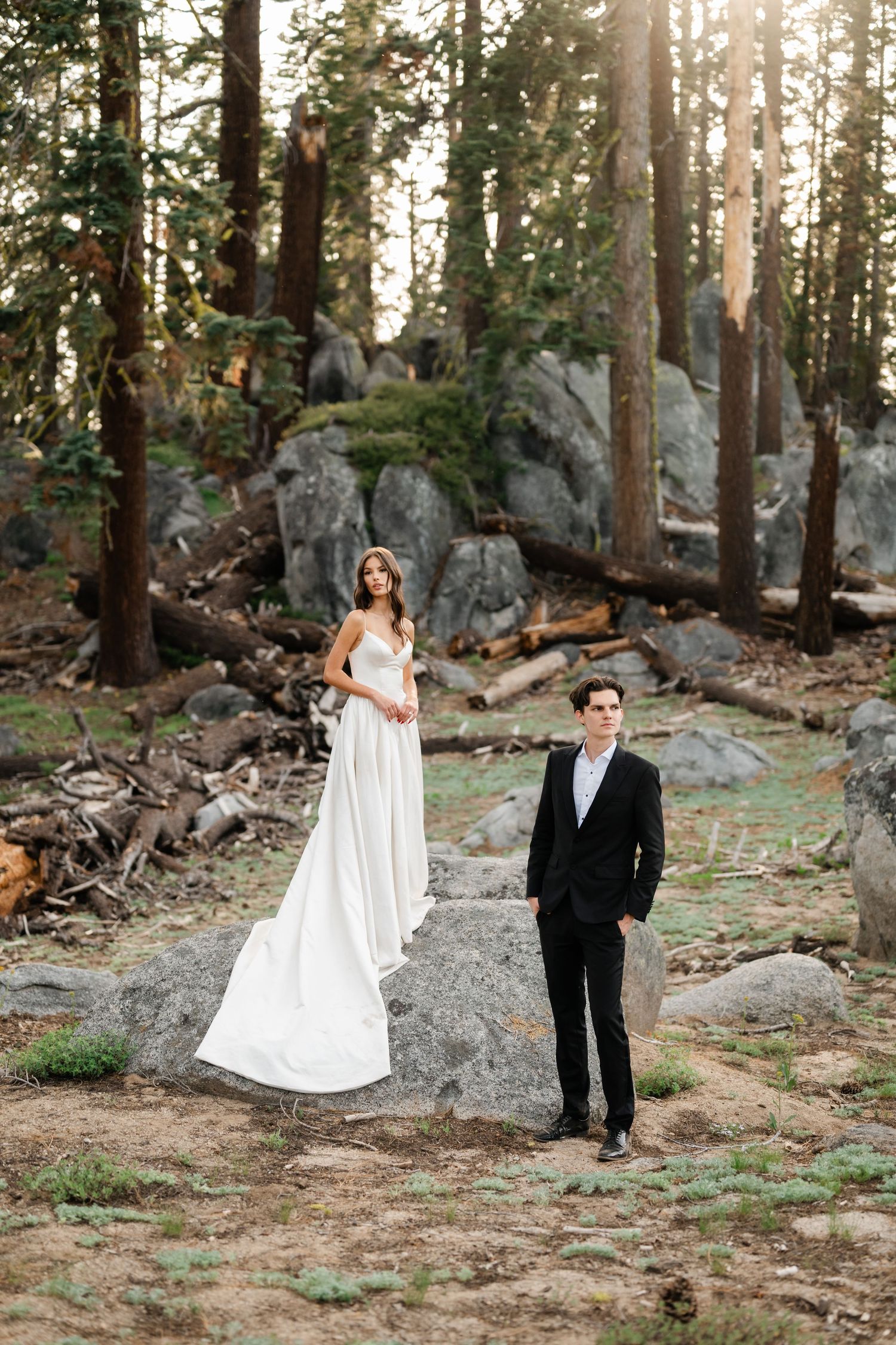 Bride in white gown and groom in black suit pose among tall pines and granite rocks at a forest wedding.