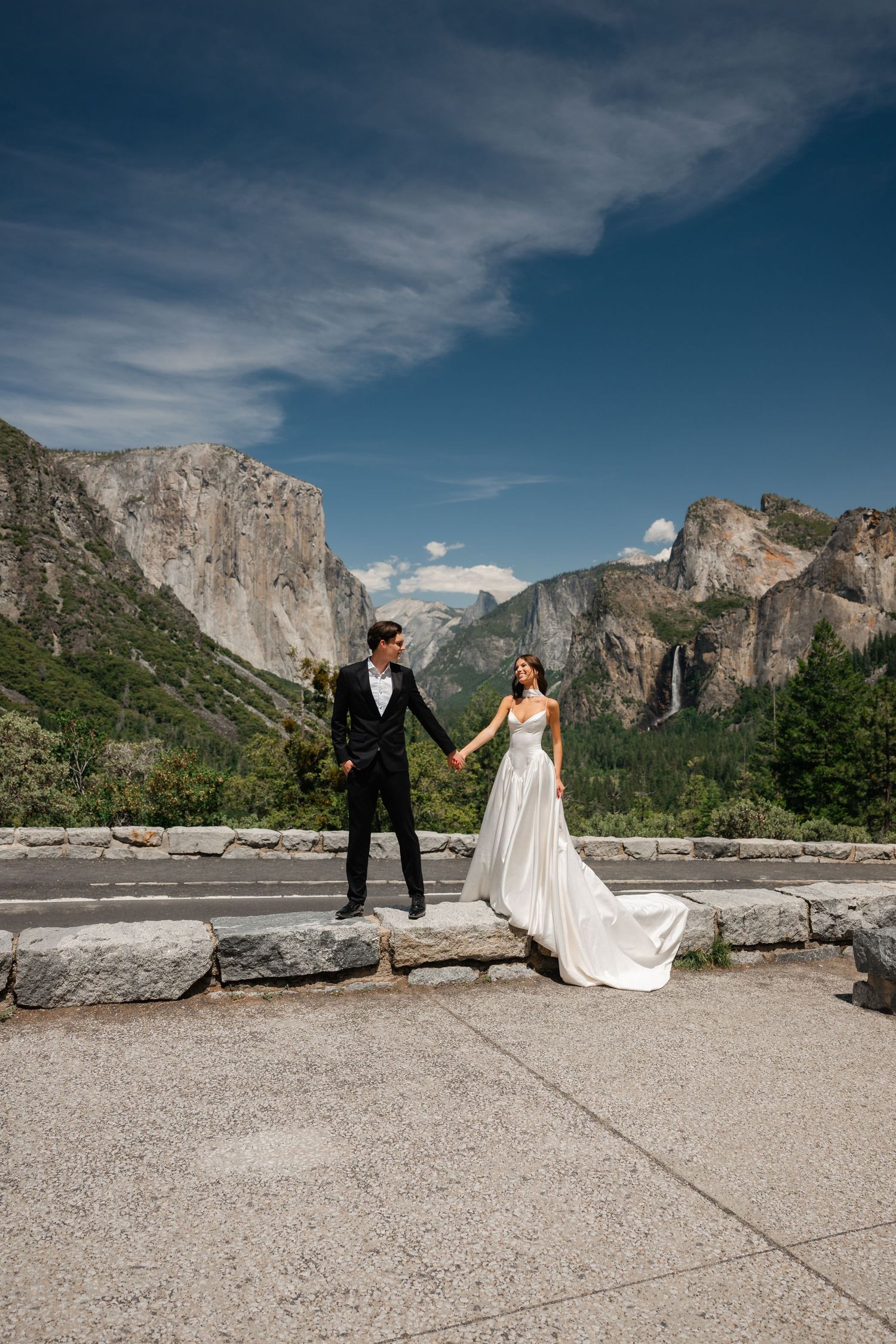 Bride and groom hold hands at Yosemite Valley overlook with El Capitan and Half Dome in the background.