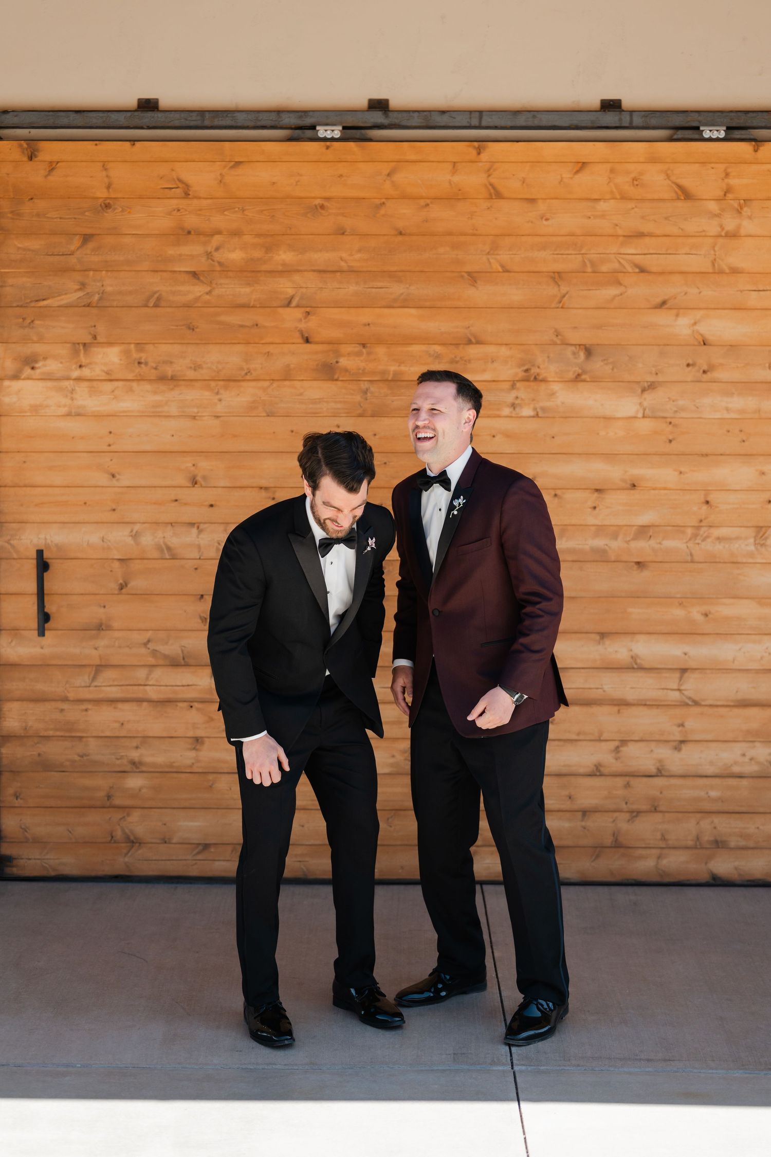 Two grooms laughing together in formal wear, one in black tuxedo and one in burgundy suit, standing before wooden barn doors.