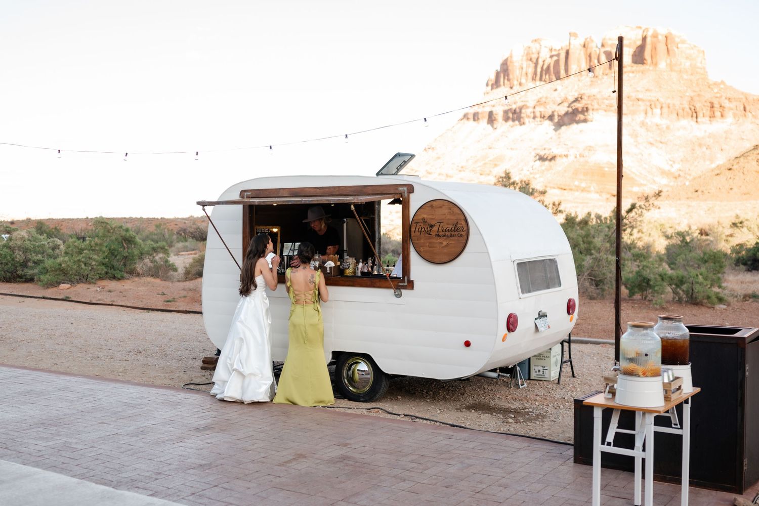 Bride and bridesmaid at a vintage white food truck bar with desert red rock backdrop at a scenic outdoor wedding.