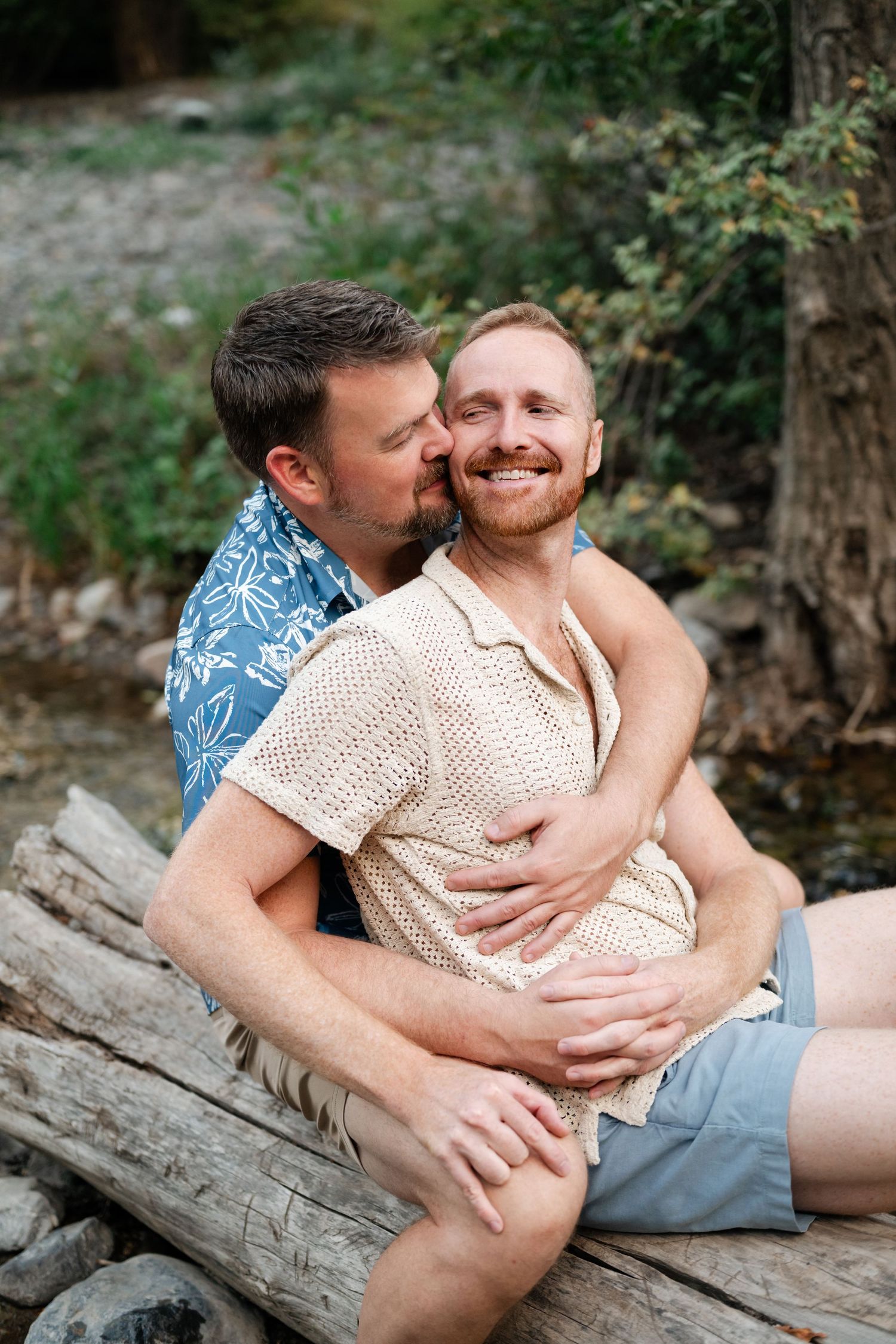 LGBTQ friendly photographer. Two men share a tender embrace on logs by a creek, one kissing the other's cheek as he smiles joyfully.