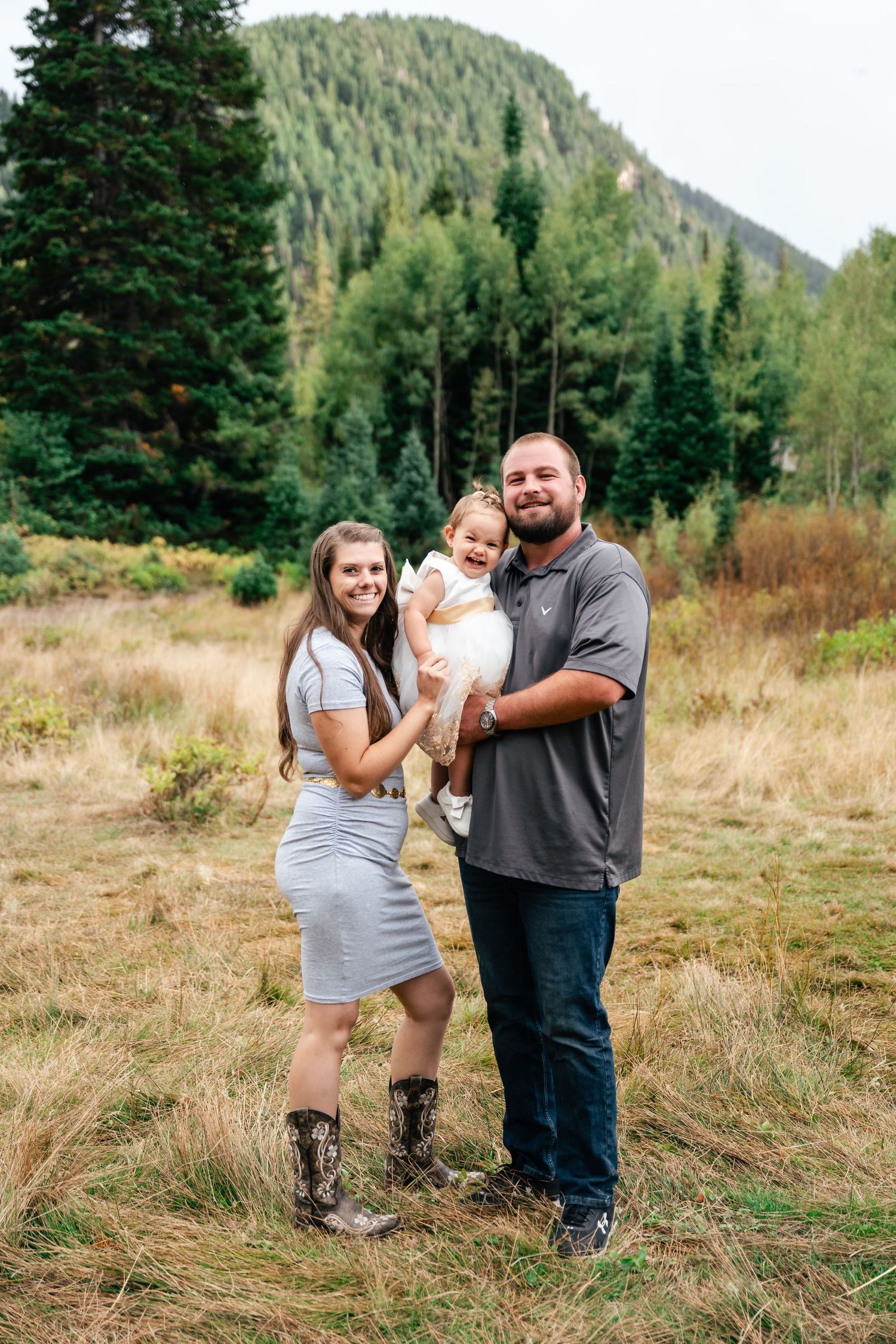 Family stands together in mountain valley with evergreen-covered slopes and autumn grass.