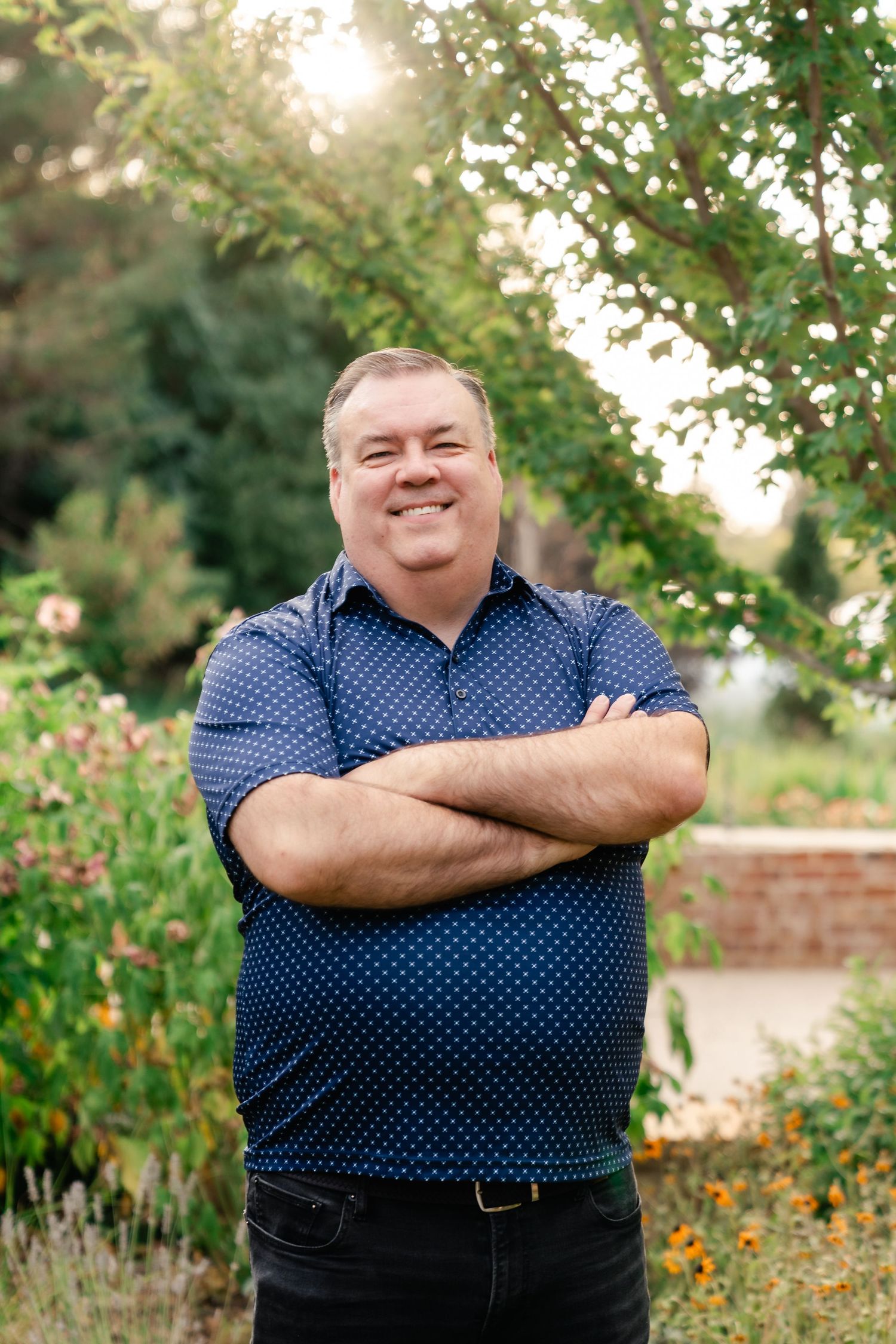 A person in a polka dot shirt poses in a garden setting with flowers and greenery in the background.