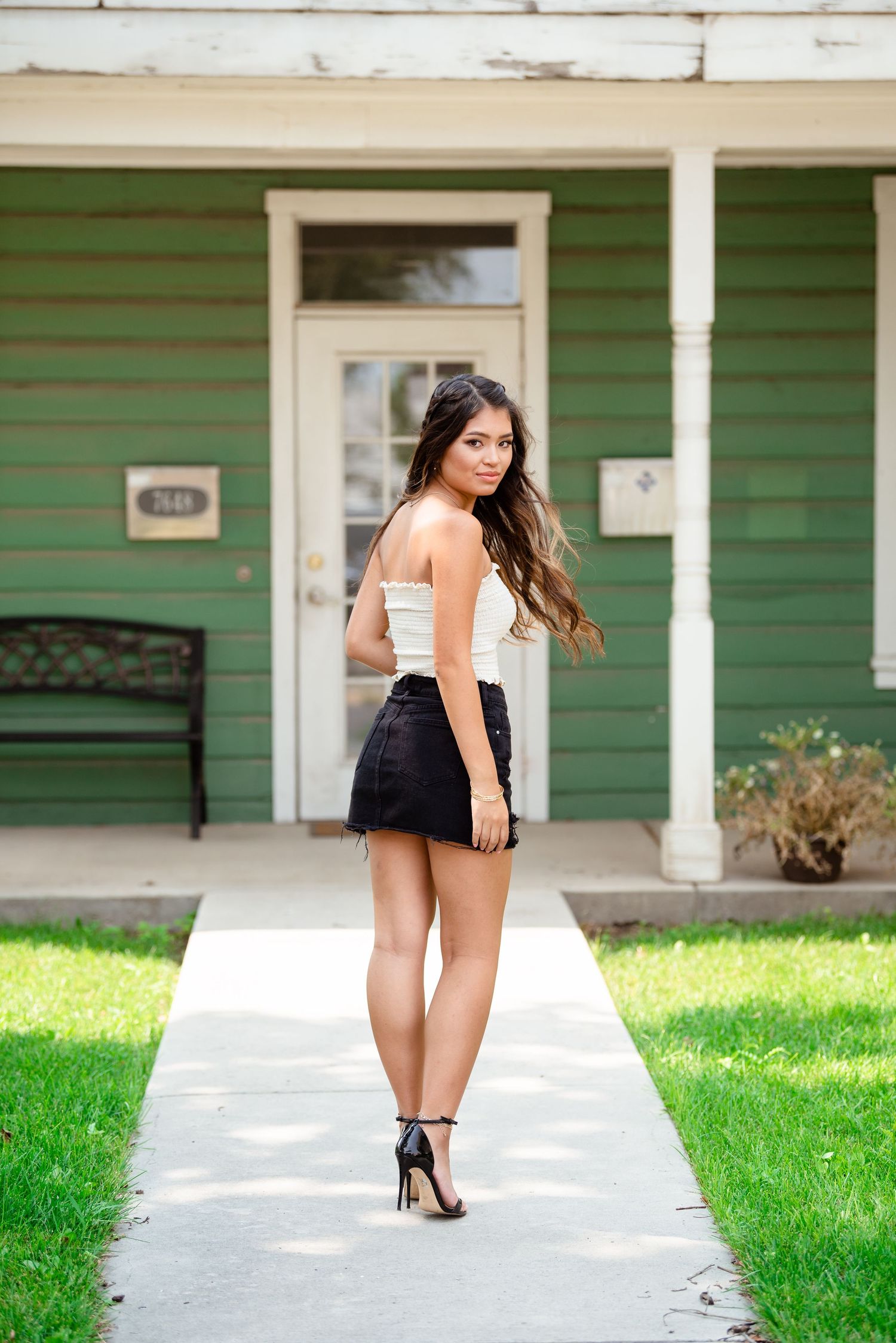 A person in a black dress poses on a concrete walkway in front of a green house with white trim.