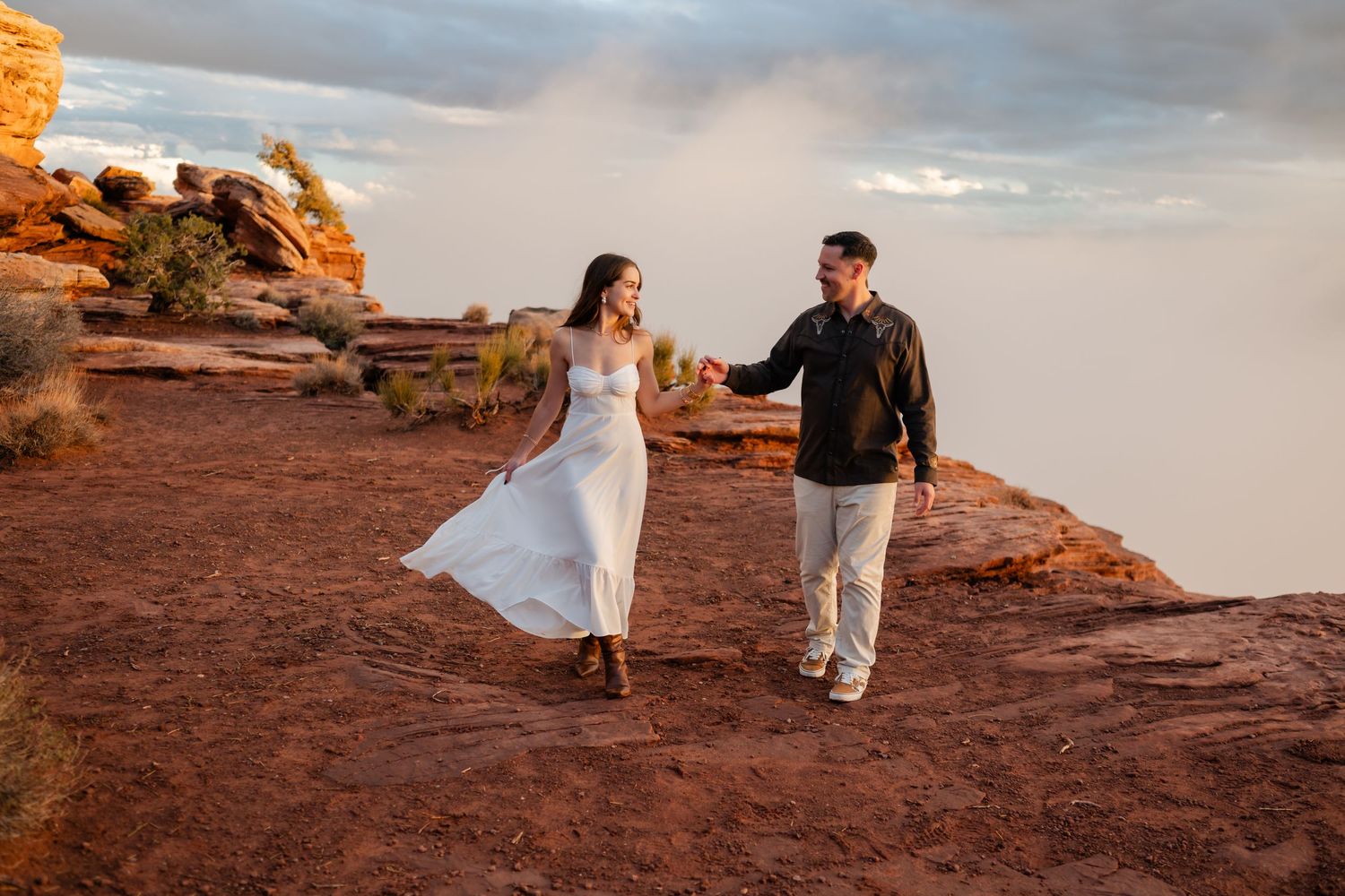 Couple in wedding attire walking along red rock formations at sunset in a desert landscape.