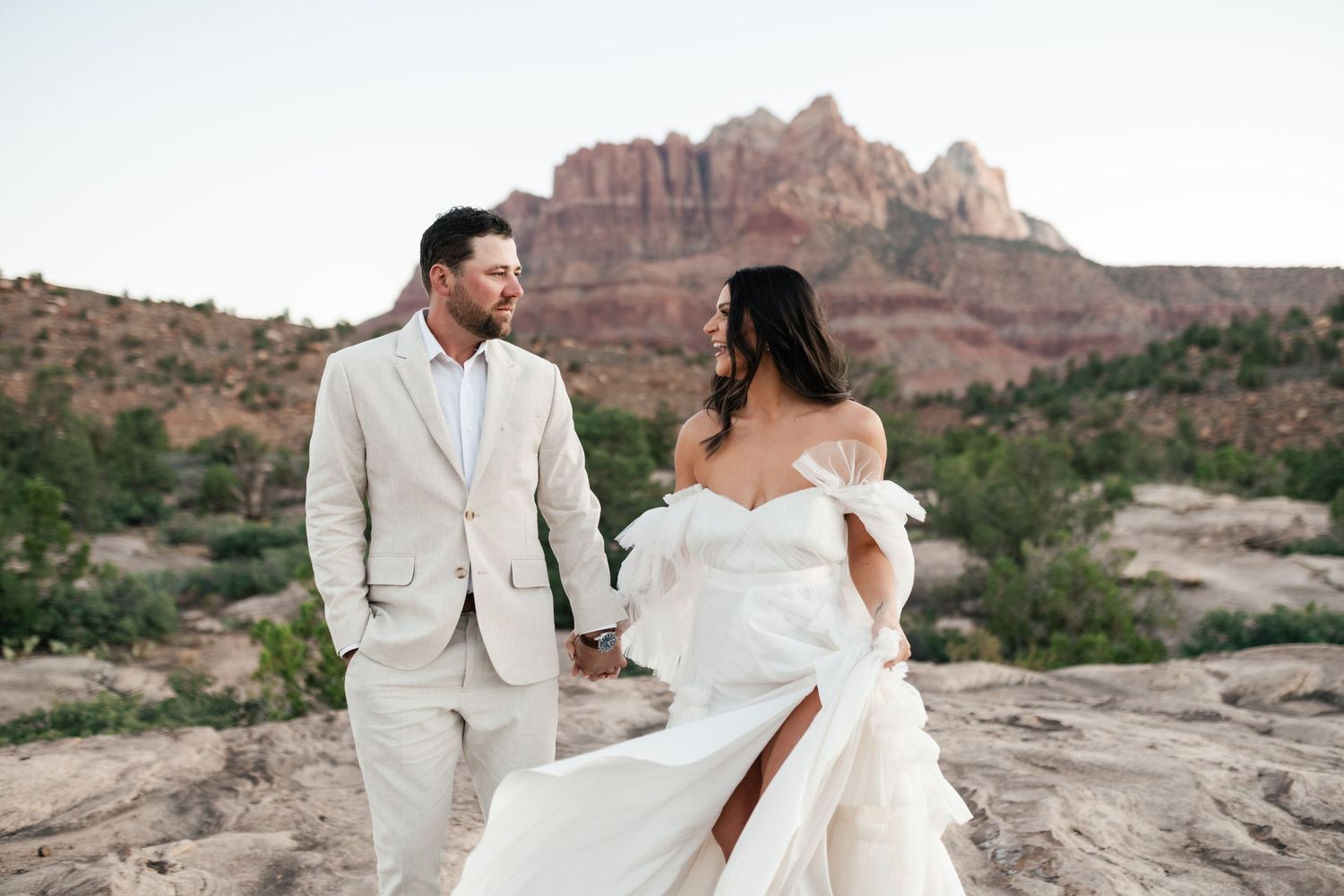 Zion Adventure. A couple in elegant white wedding attire poses together against the stunning red rock formations of a desert landscape.