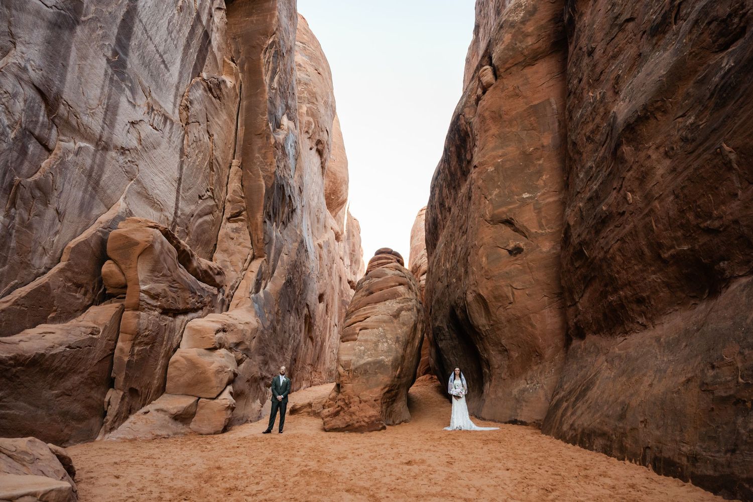 Hikers explore towering red rock canyon walls in Arches National Park during golden hour.