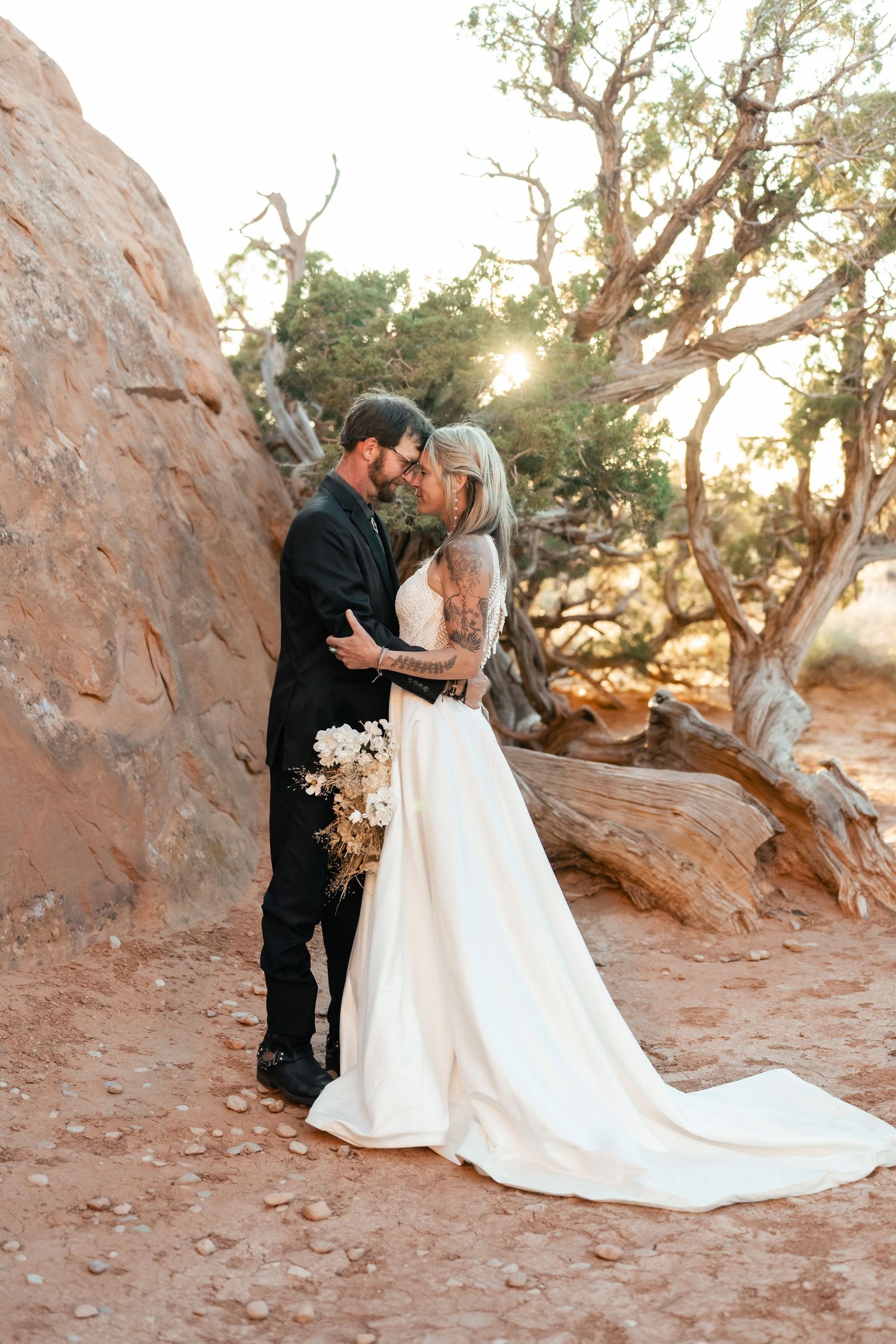 A couple in wedding attire share romantic moments at sunset among desert rocks and twisted trees.