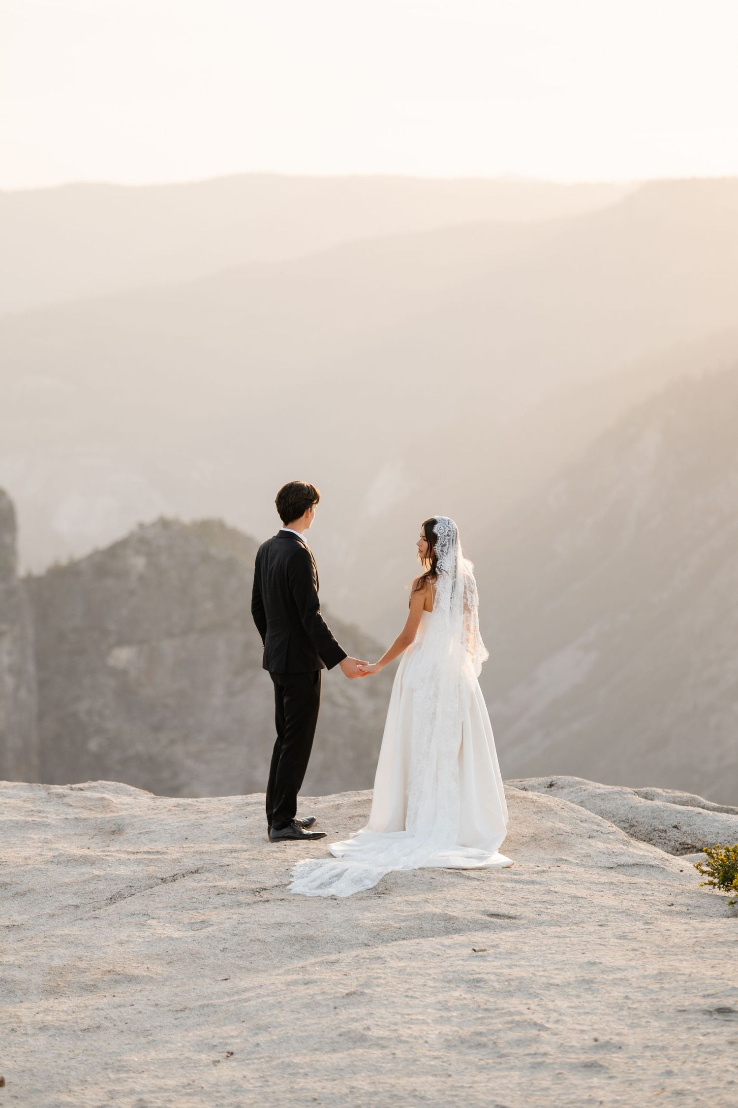 Bride and groom hold hands facing each other on rocky mountain overlook at sunset.