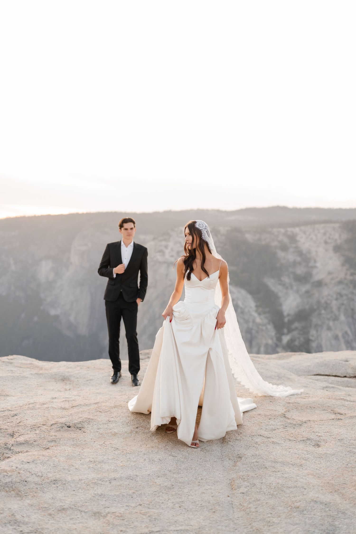 Bride in elegant white gown with train walks on rocky cliff edge as groom approaches in black suit.