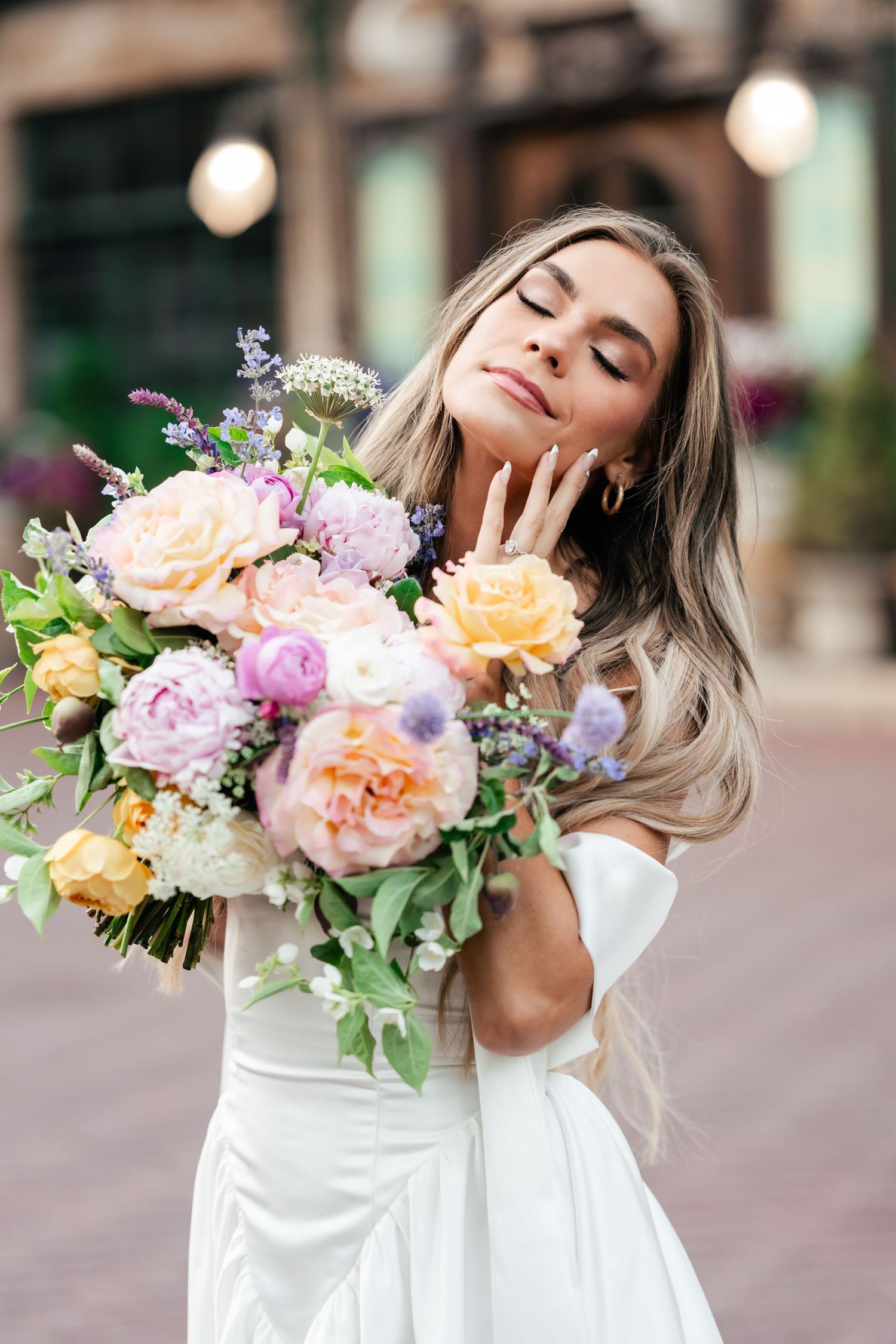 Person in white dress holds luxurious bouquet of peach, pink and purple flowers with eyes closed outside.
