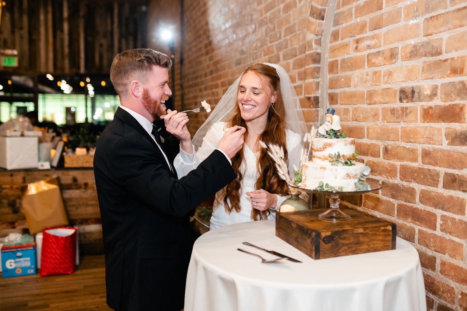 Newlyweds share a playful moment while feeding each other wedding cake in front of a rustic brick wall.