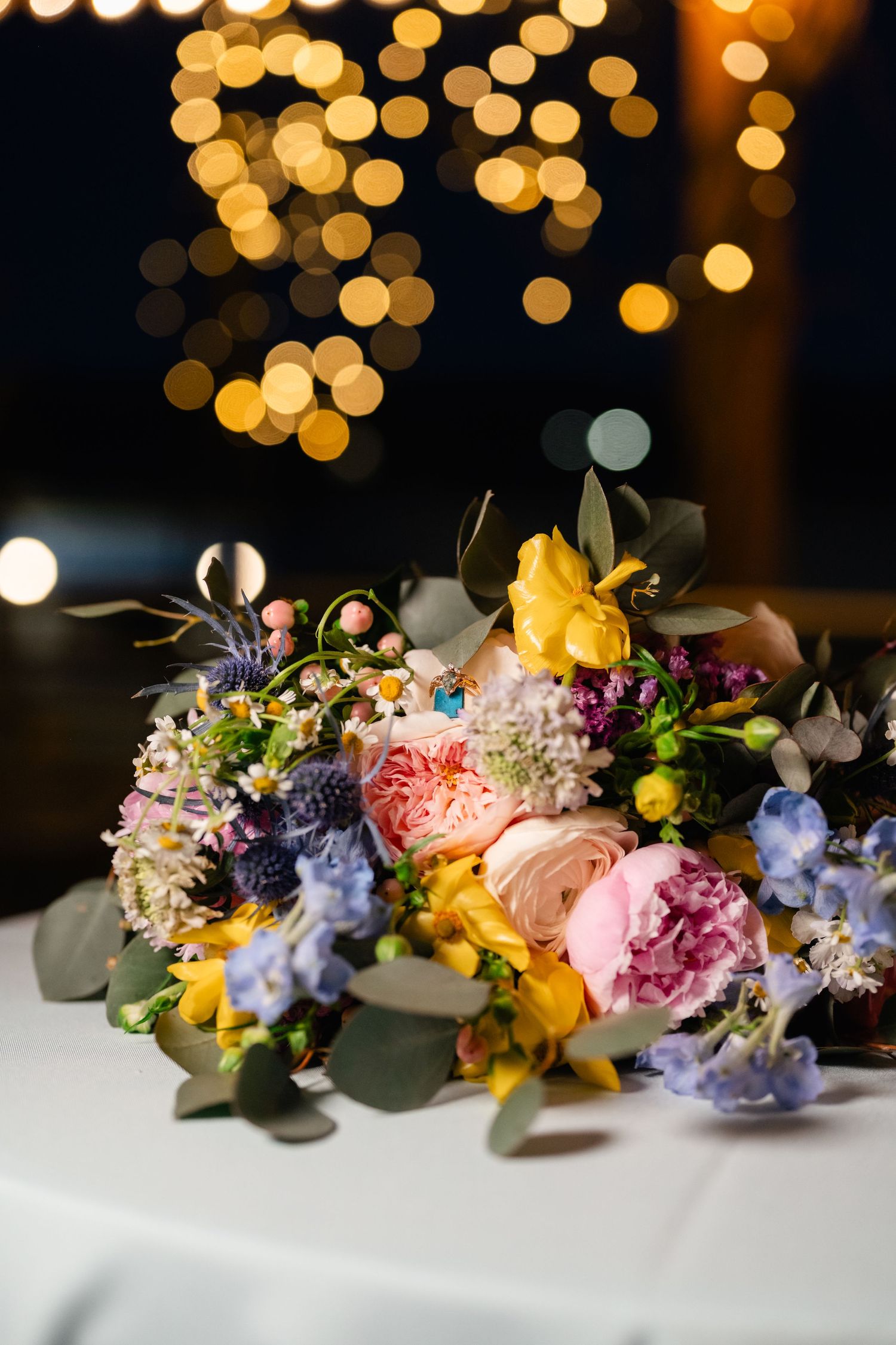 A colorful wedding centerpiece with pink and blue flowers sits on a white table beneath glowing bokeh lights.