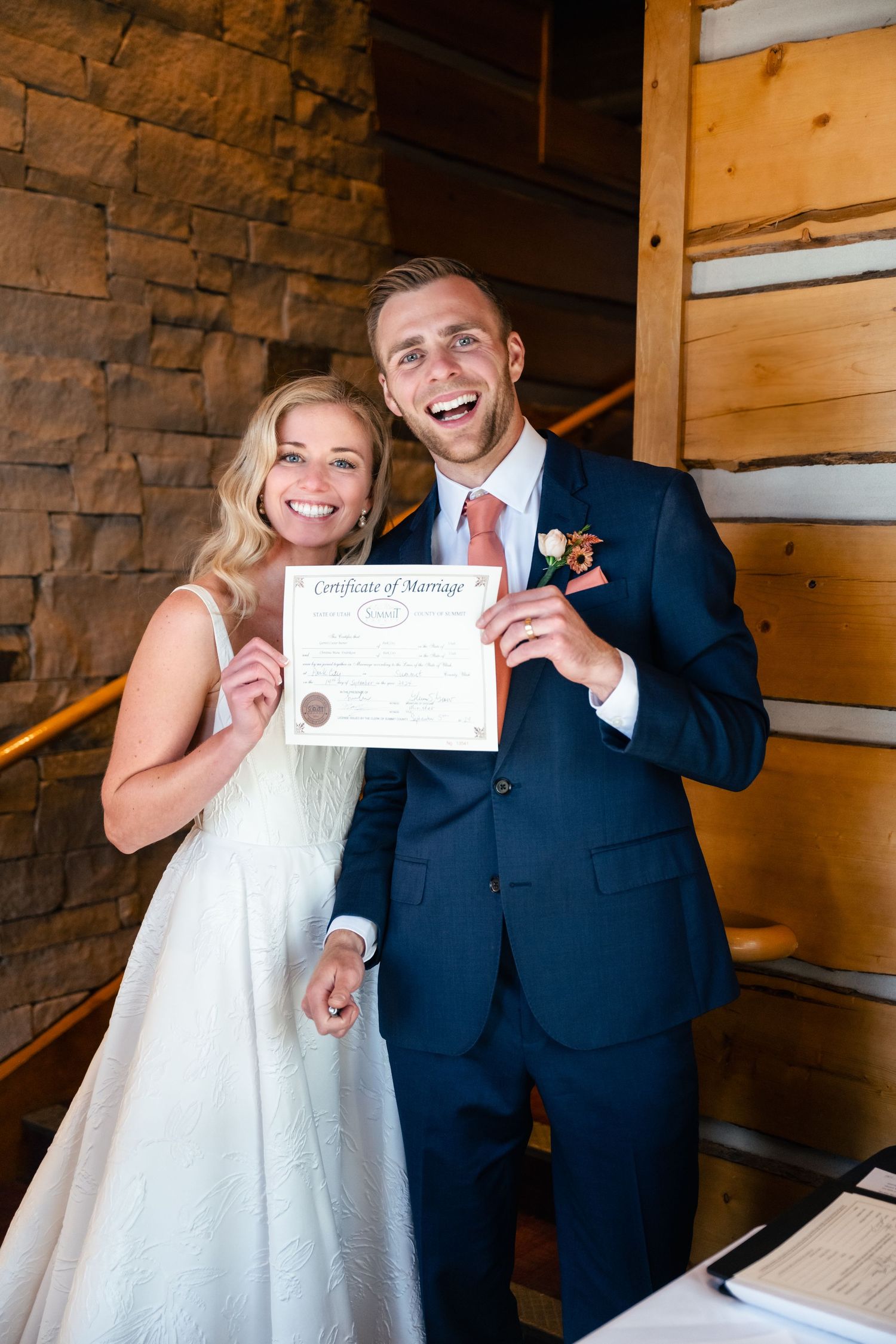 A joyful couple in wedding attire show off their marriage certificate while posing in a rustic wooden venue. Park City Wedding Photographer.