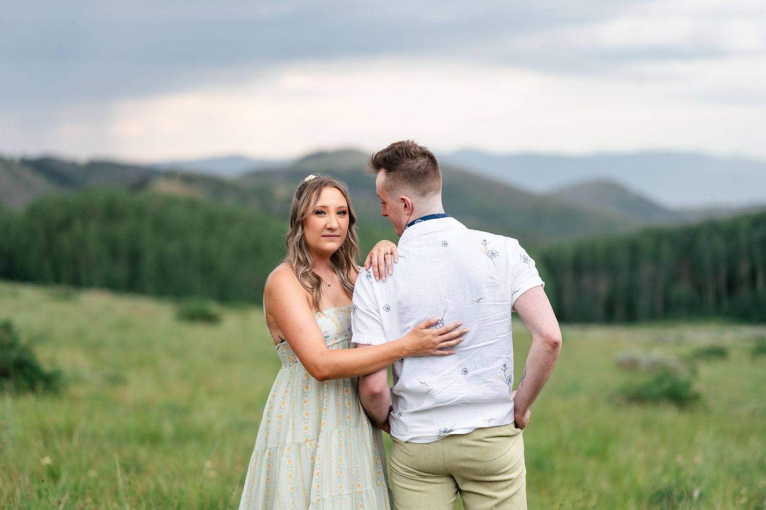 A romantic moment captured between a couple in light summer attire against a mountainous landscape. Utah mountain engagement session