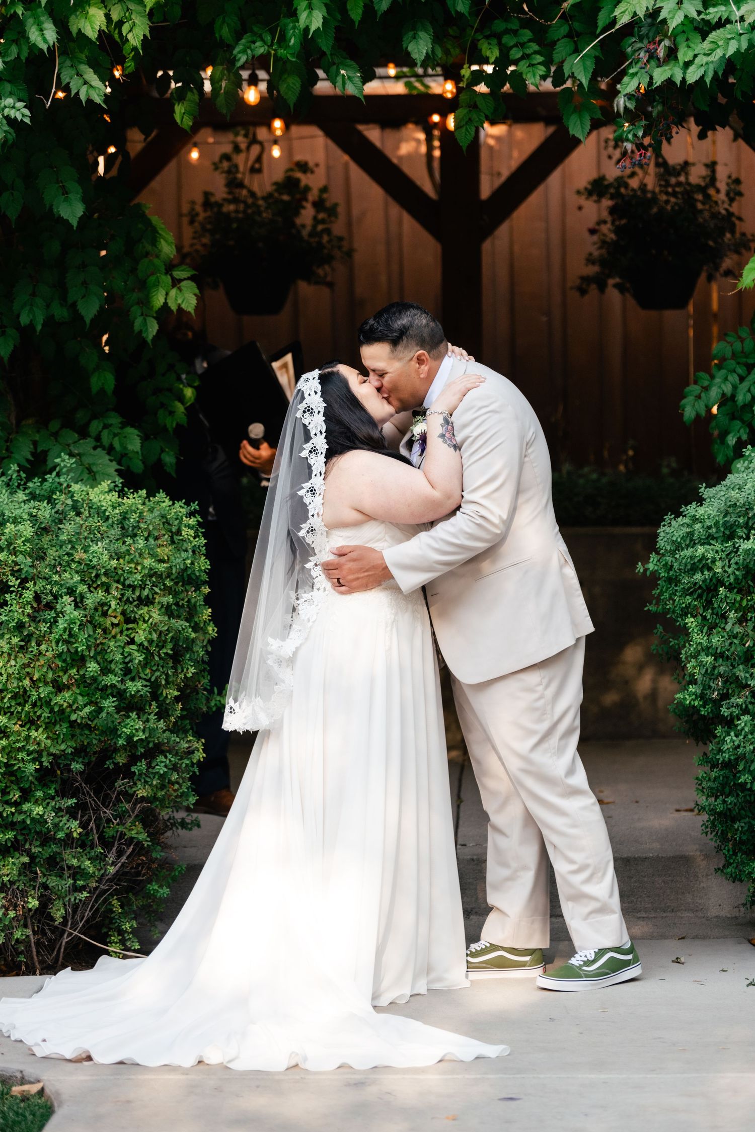 Romantic outdoor wedding portrait in front of wooden archway decorated with hanging plants and greenery.