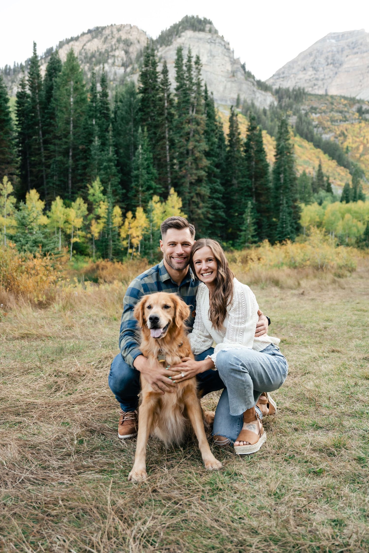 A couple poses with their golden retriever against stunning mountain scenery with fall colors.