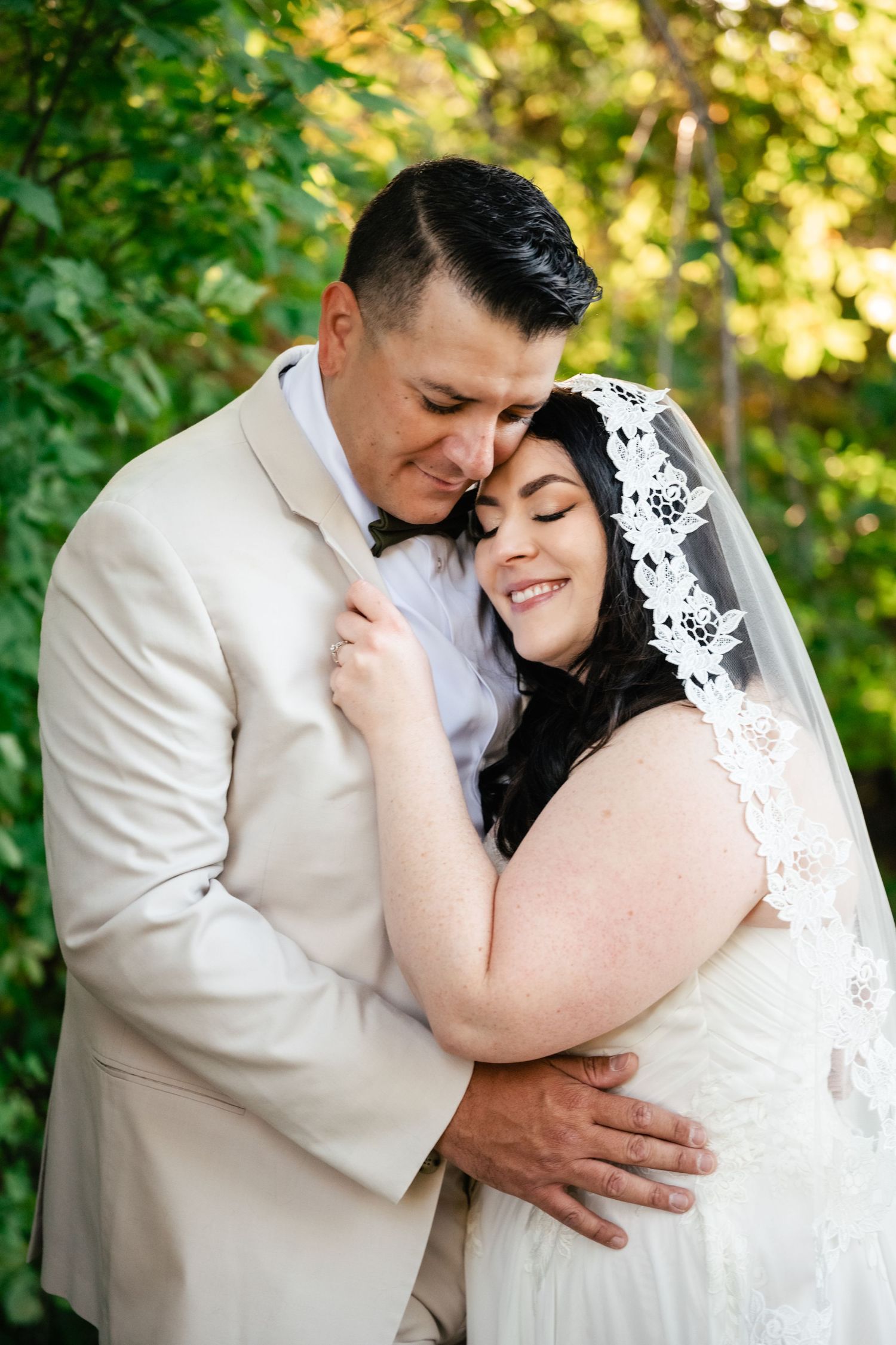 A romantic outdoor wedding portrait shows a couple embracing while the bride wears a delicate lace veil.