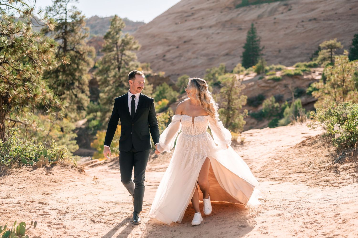 A couple walks hand in hand along a desert path wearing formal wedding attire.