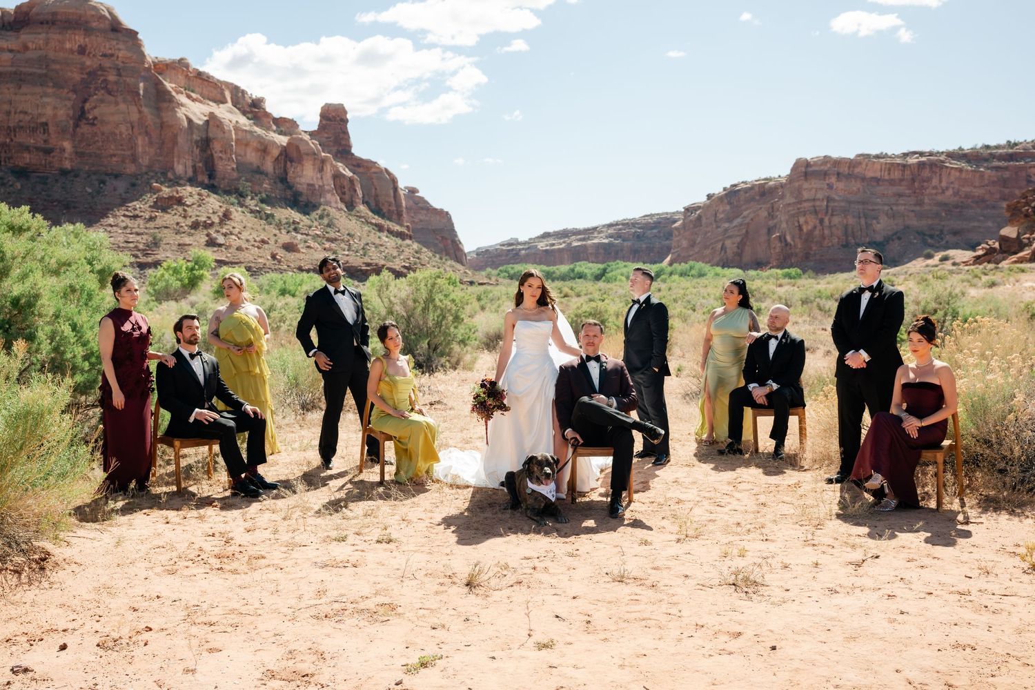 Wedding party poses in red rock canyon desert landscape with bride, groom, groomsmen in black, bridesmaids in burgundy.