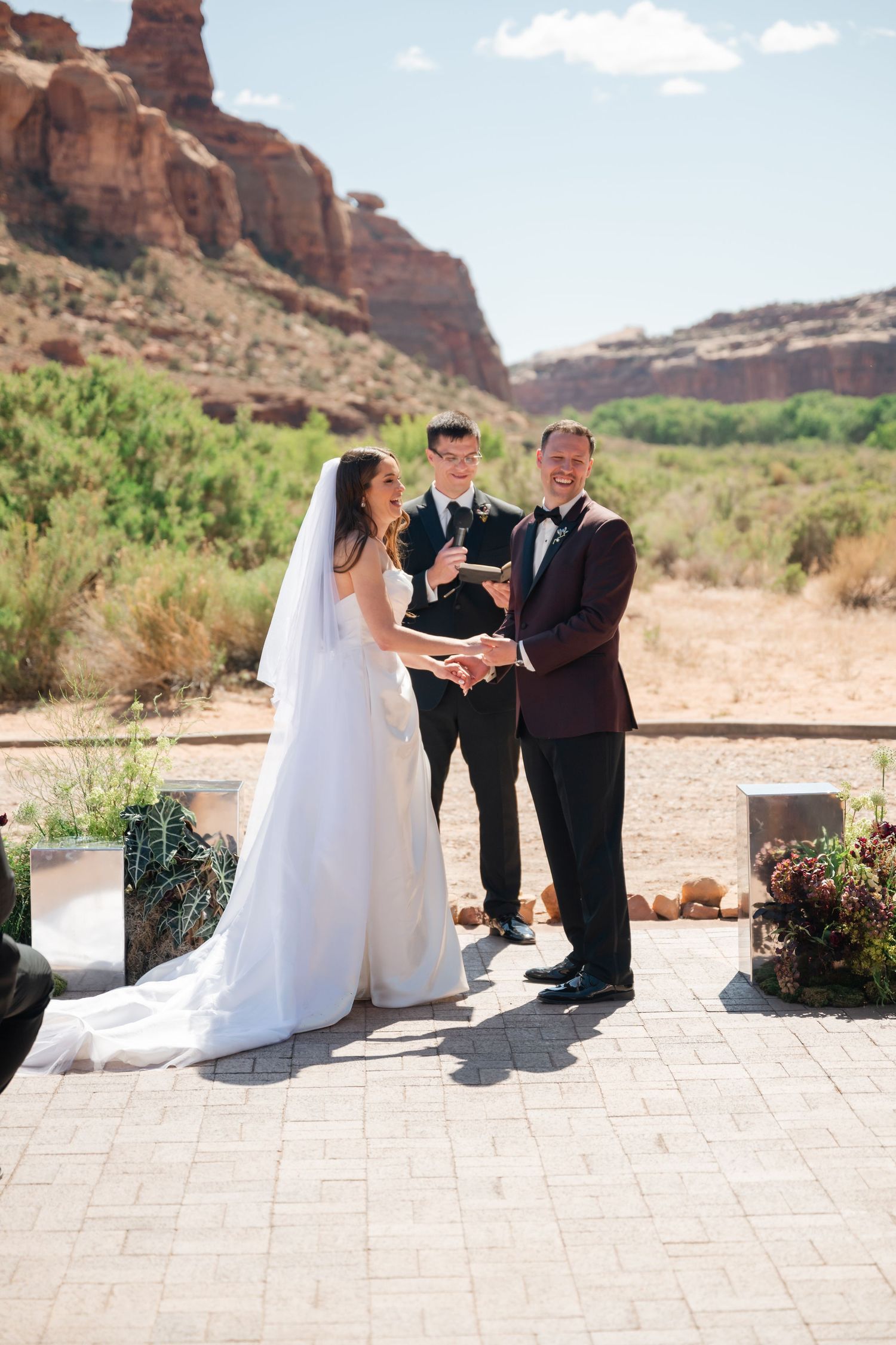 Bride and groom exchange vows outdoors with red rock canyon backdrop during sunny desert wedding ceremony.