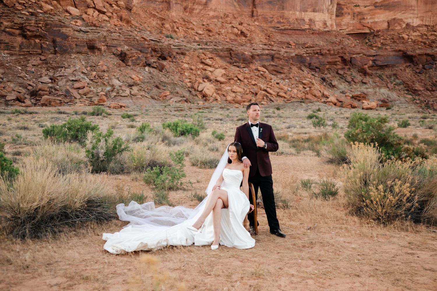 Bride in white gown and groom in black tuxedo pose before red rock canyon desert landscape.