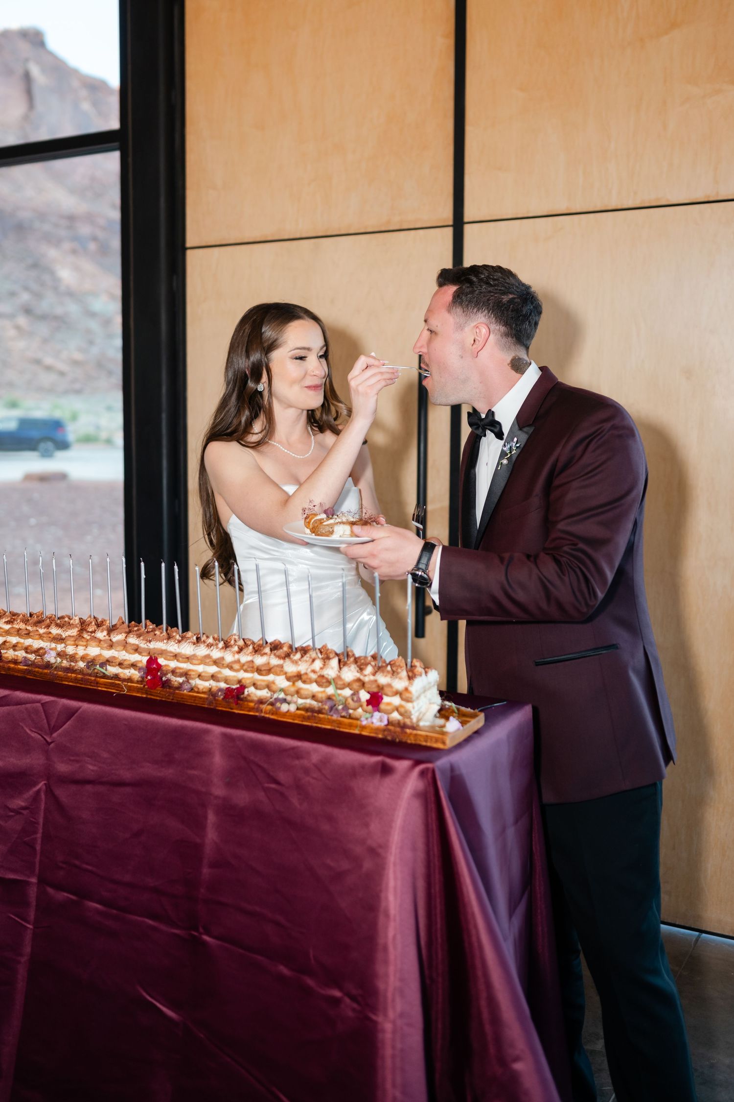 Bride feeds groom wedding cake at elegant desert venue reception table with burgundy linen.
