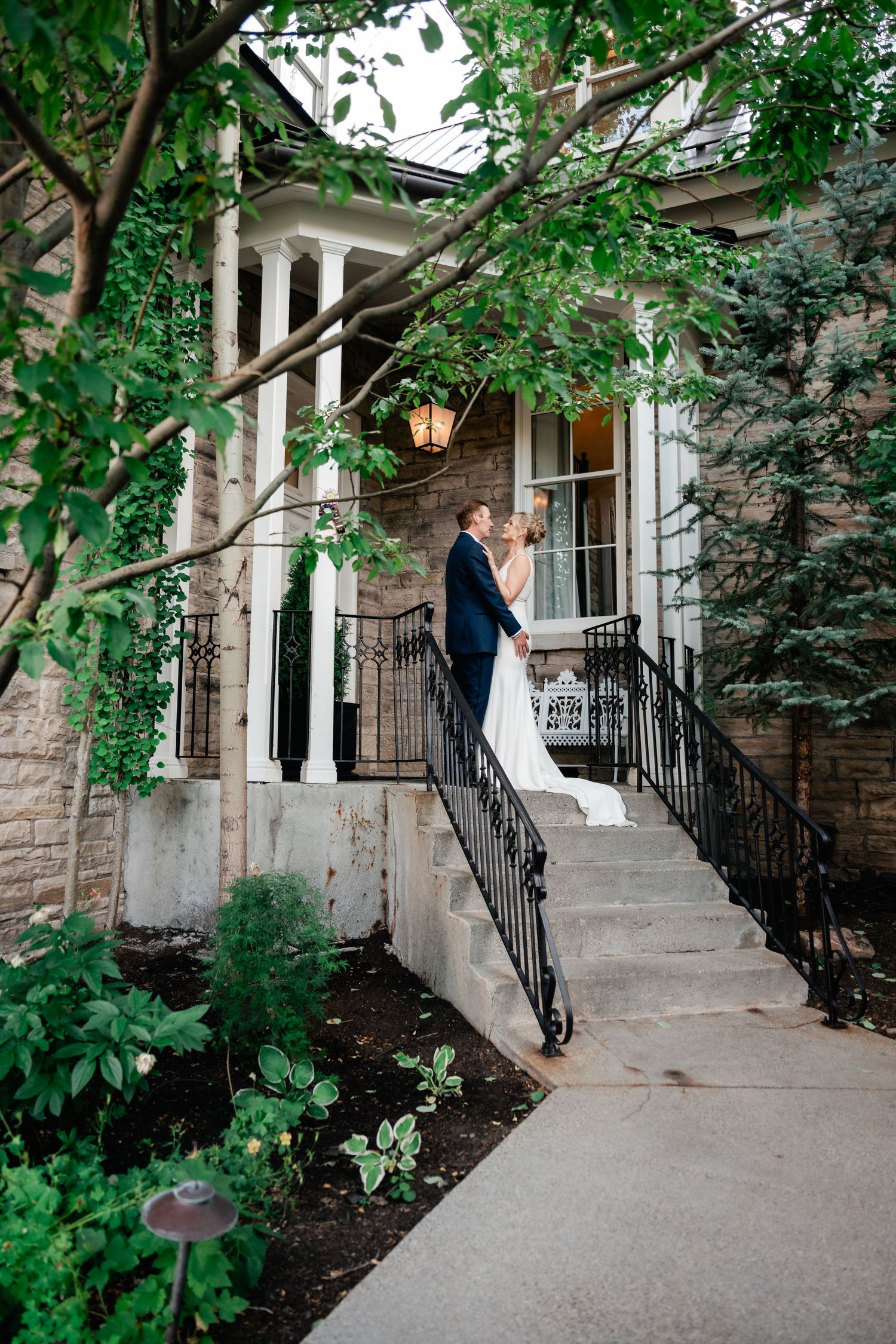 Wedding couple shares intimate moments along a stone building exterior with black railings and green foliage.