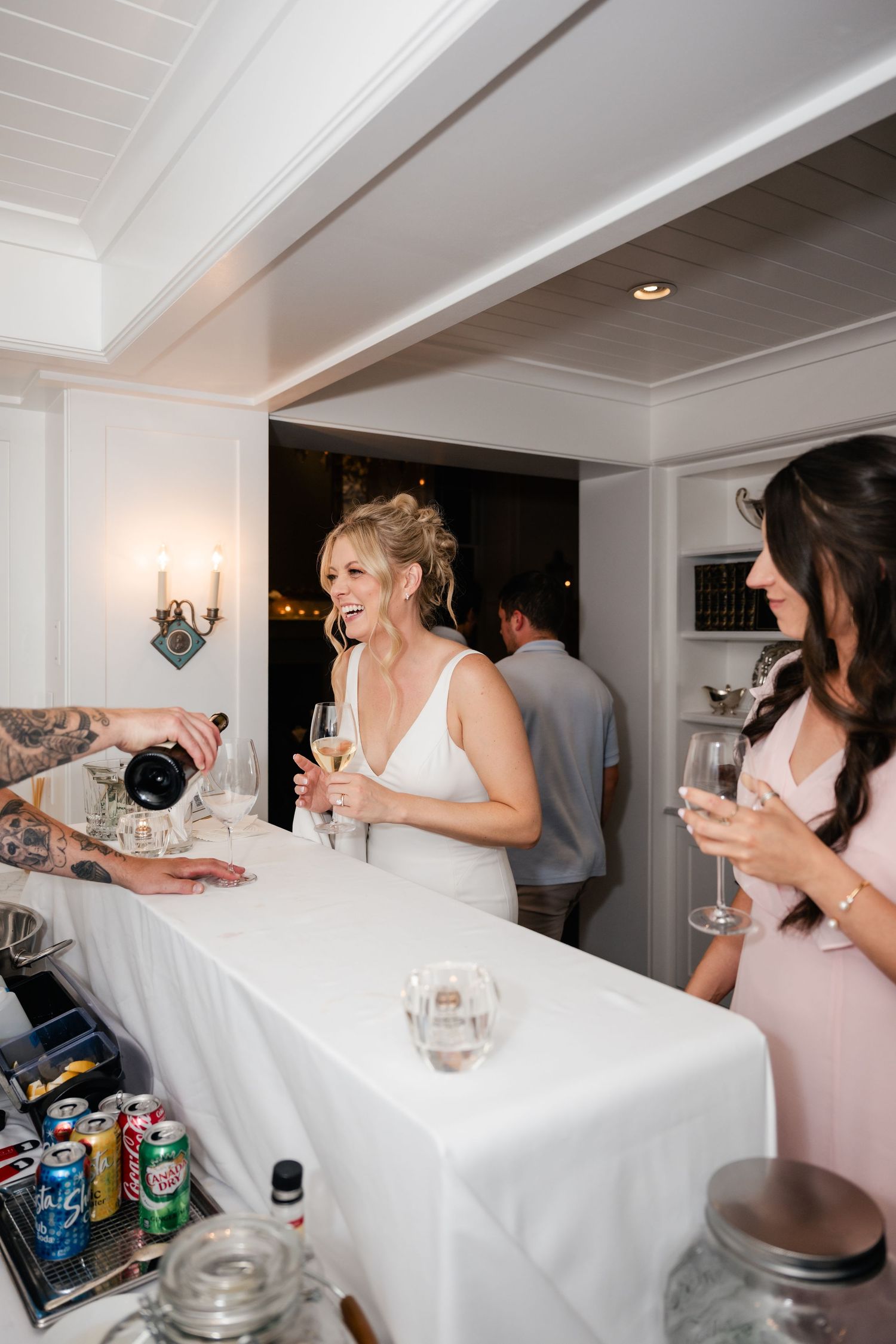 Wedding party members laugh and socialize around a white-covered table at an elegant indoor reception venue.