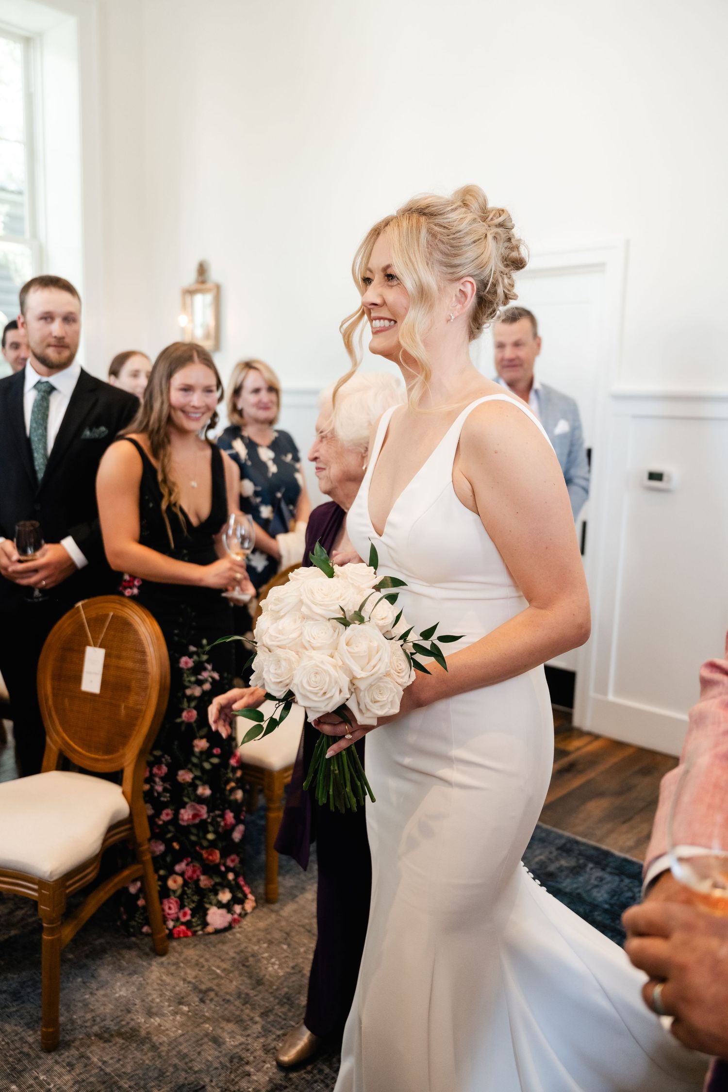 Wedding guests gather in a bright ceremony space with white walls and wood flooring.