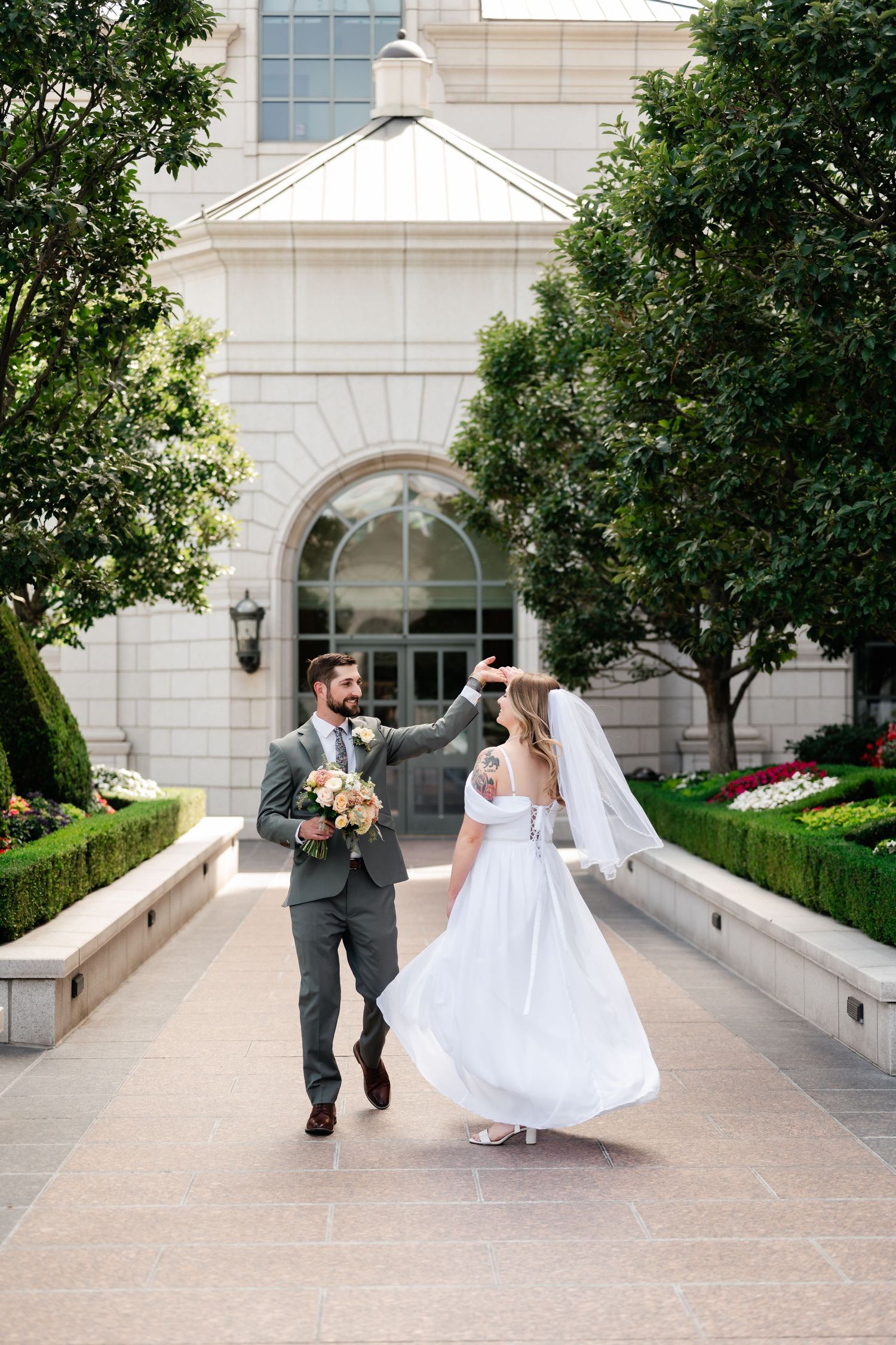 A couple walks along a garden path with greenery and a grand stone building entrance in the background.