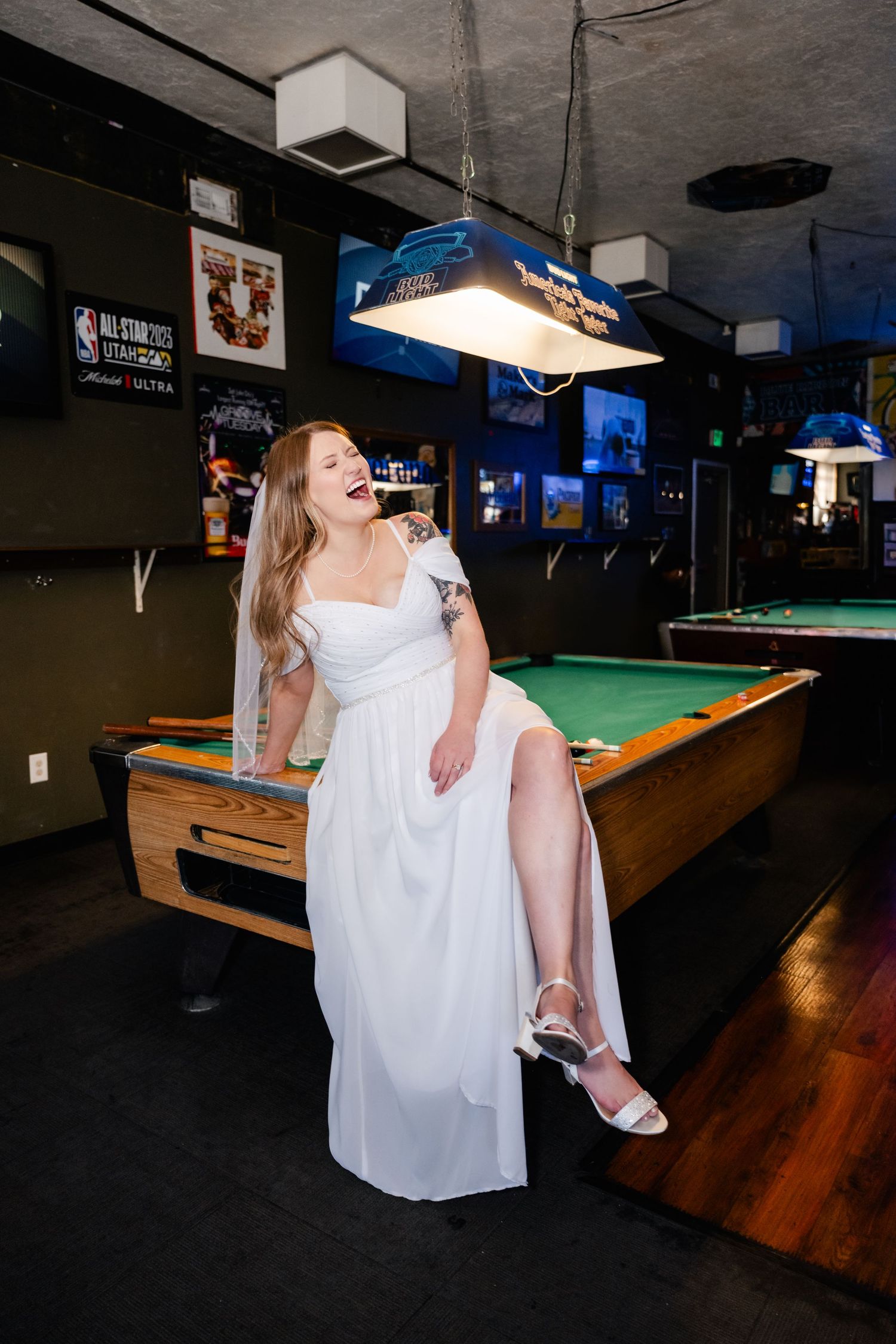 A bride in a white dress poses playfully on a pool table in a sports bar setting.