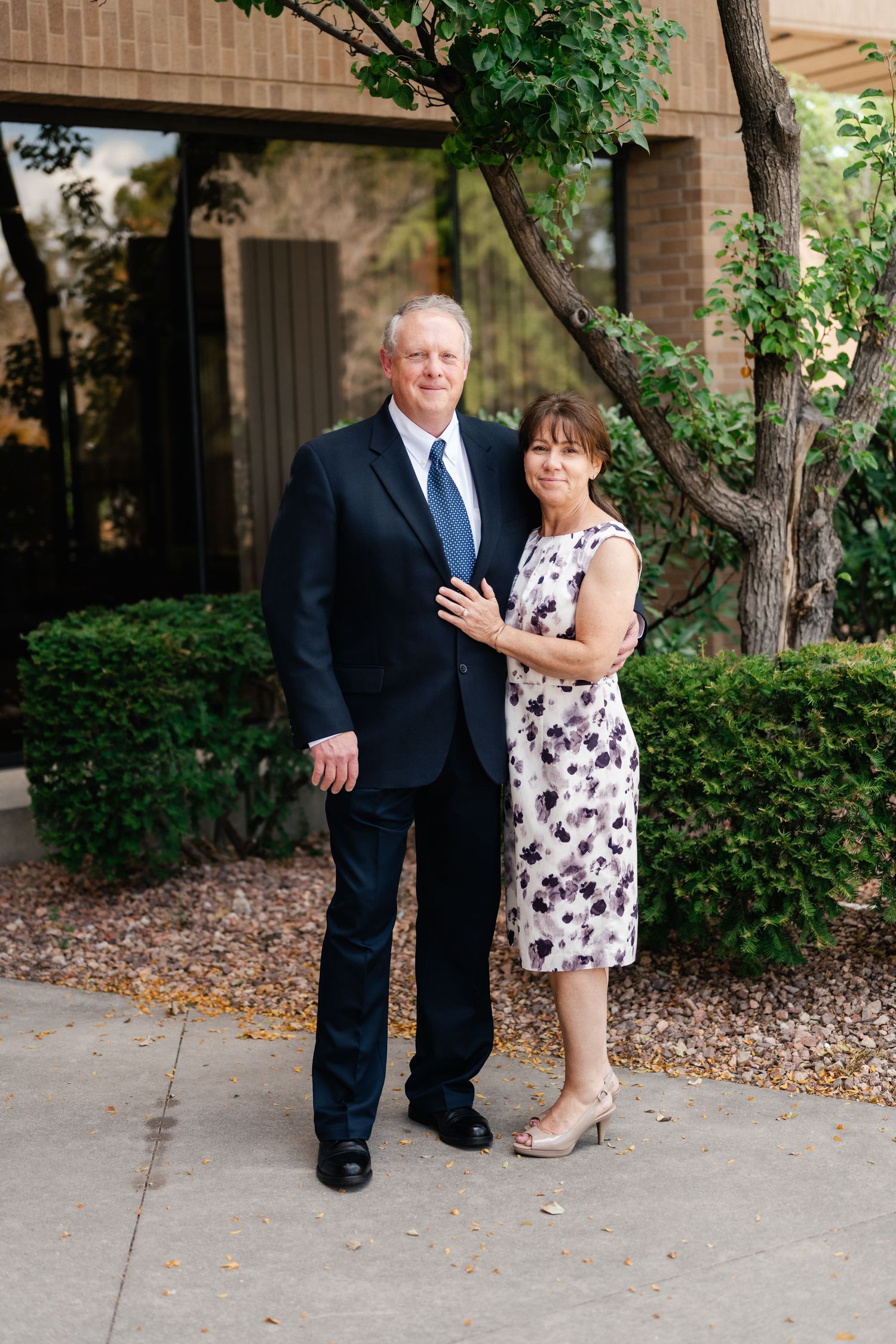 A couple poses together outdoors in formal attire near landscaped bushes and a brick building.
