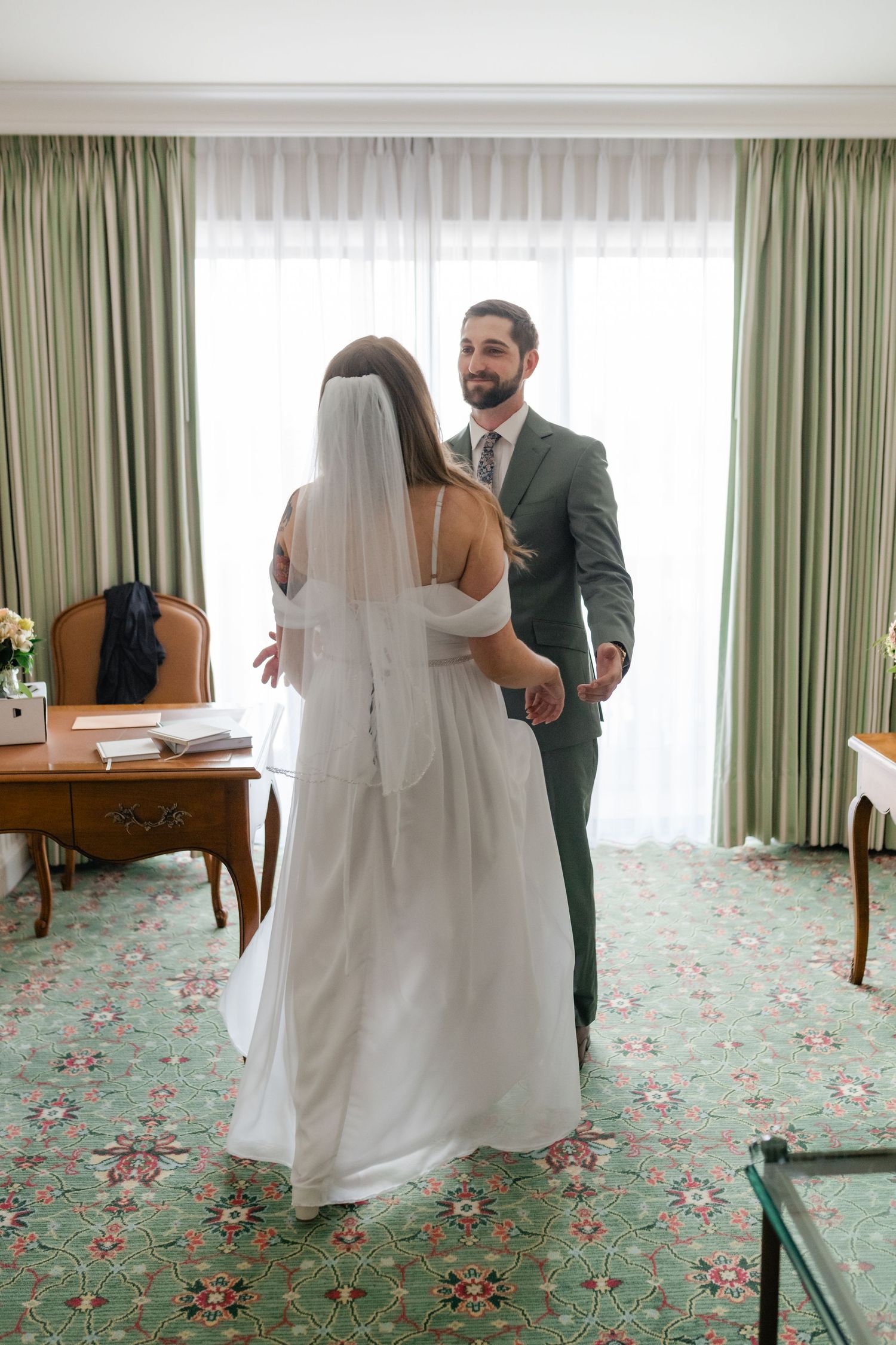 A first look wedding moment captured in a hotel room with floral carpeting and sage green drapes.